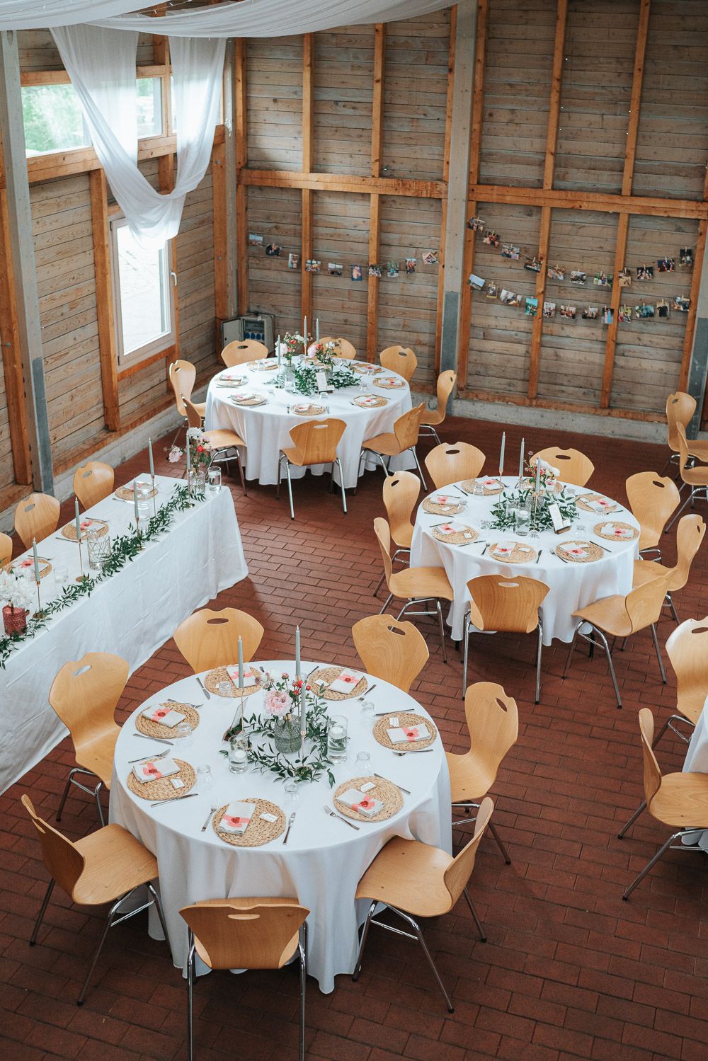 Rustic wedding reception setup with round tables, white linens, wooden chairs and string lights in a barn-style venue.