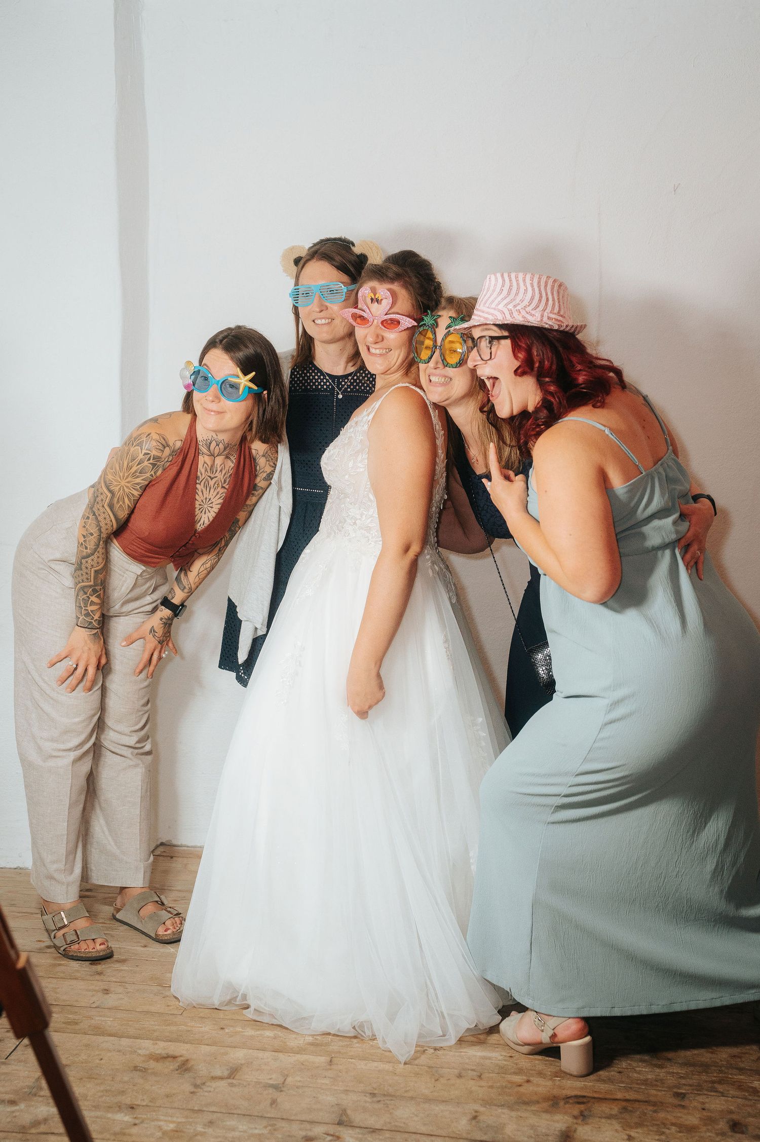 Bride in a white tulle gown poses with four bridesmaids wearing playful photo booth glasses and a pink hat at a rustic wedding.