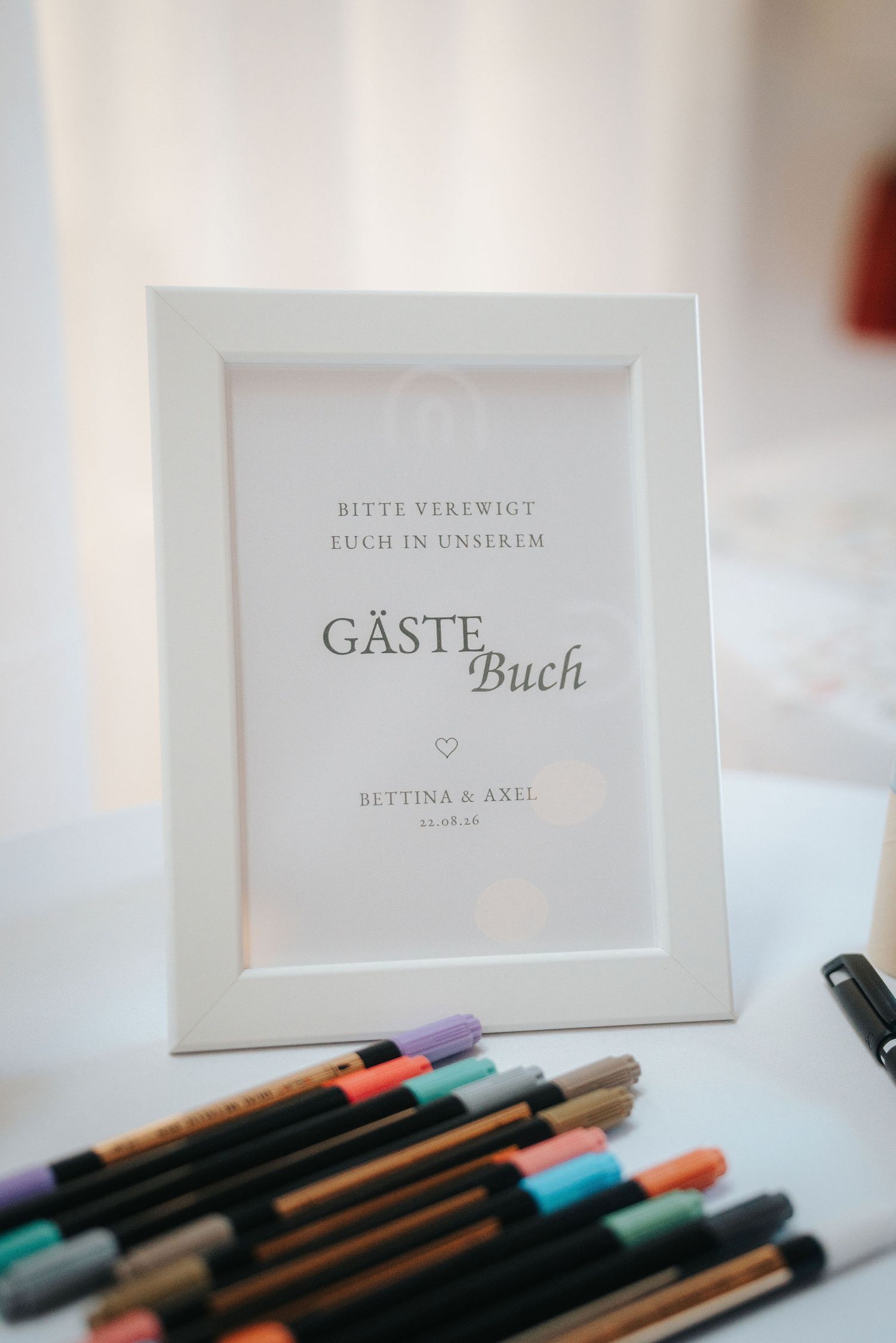 White framed German wedding guest book sign for Bettina & Axel beside a row of pastel and metallic markers on a table.