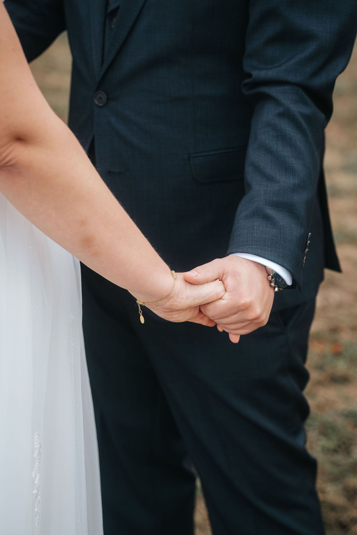 Bride in a white dress and groom in a dark pinstripe suit hold hands in a close-up wedding moment outdoors.