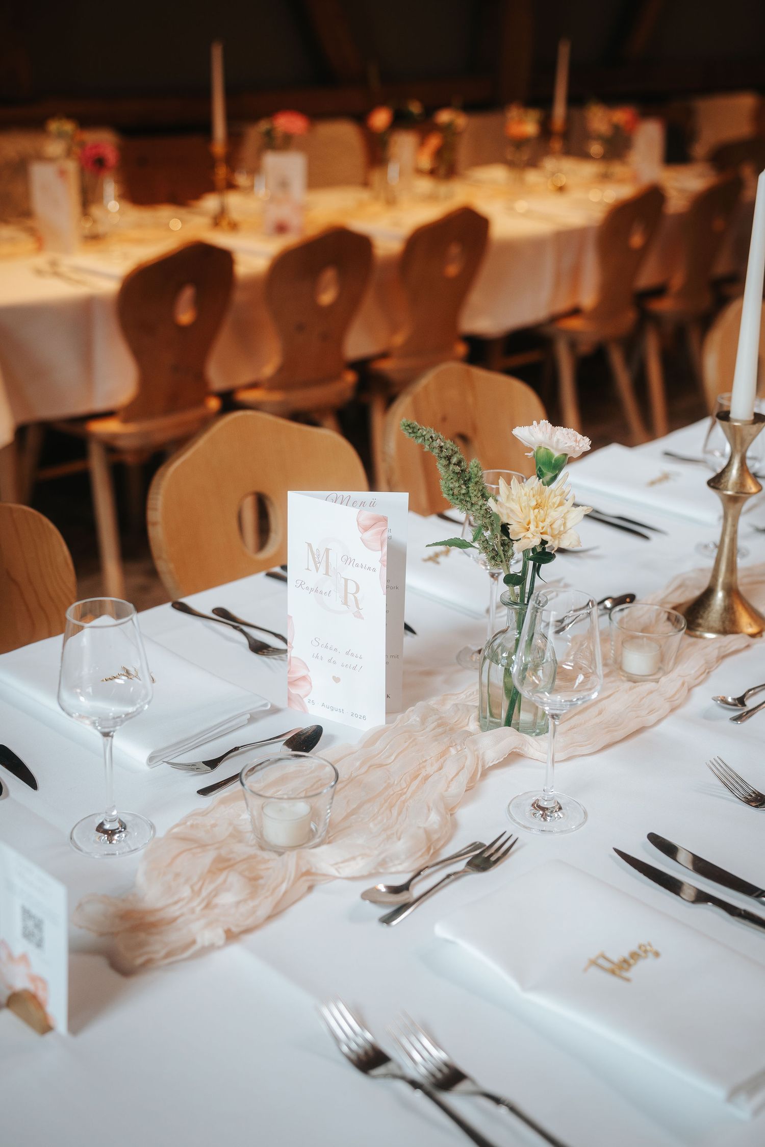 Rustic wedding reception table with white linens, folded napkins, wine glasses, a floral centerpiece, menu card and wooden chairs.