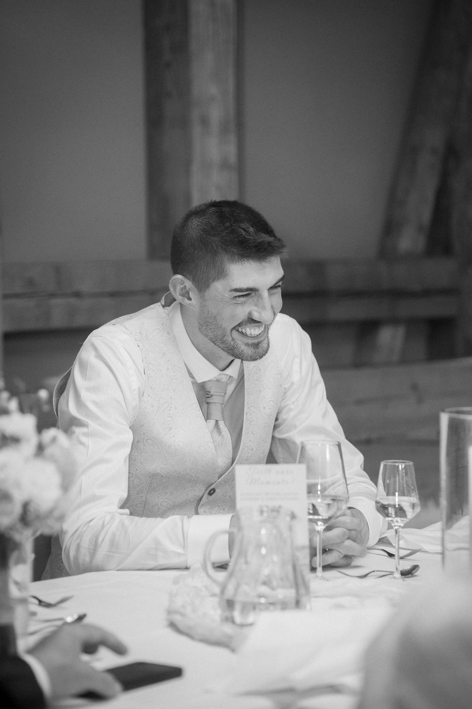 Smiling man in a vest and tie laughs at a wedding reception table set with wine glasses, a water jug and flowers in black and white.
