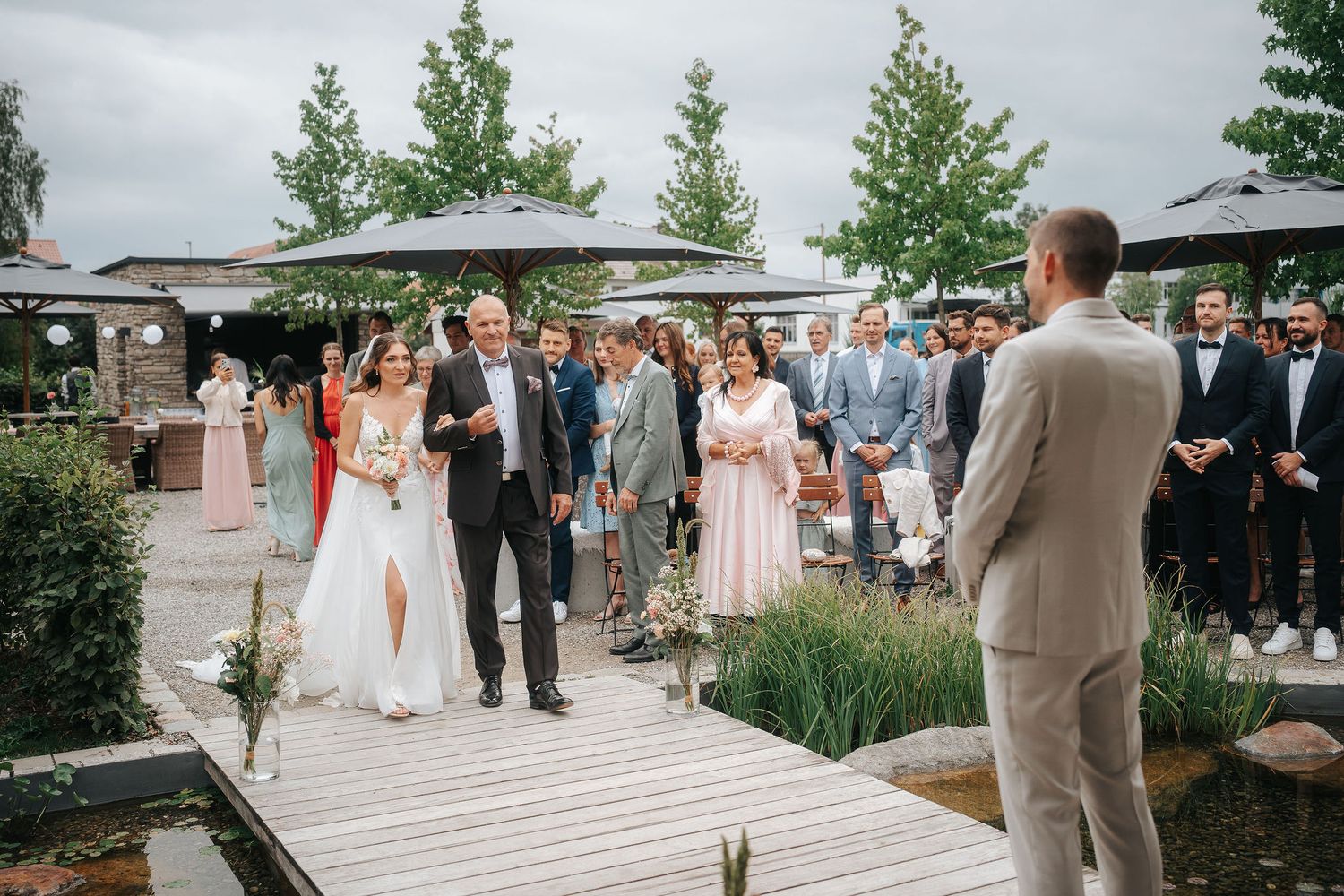 Bride in a white gown walks down a wooden boardwalk with a man in a dark suit as wedding guests watch at an outdoor garden ceremony.