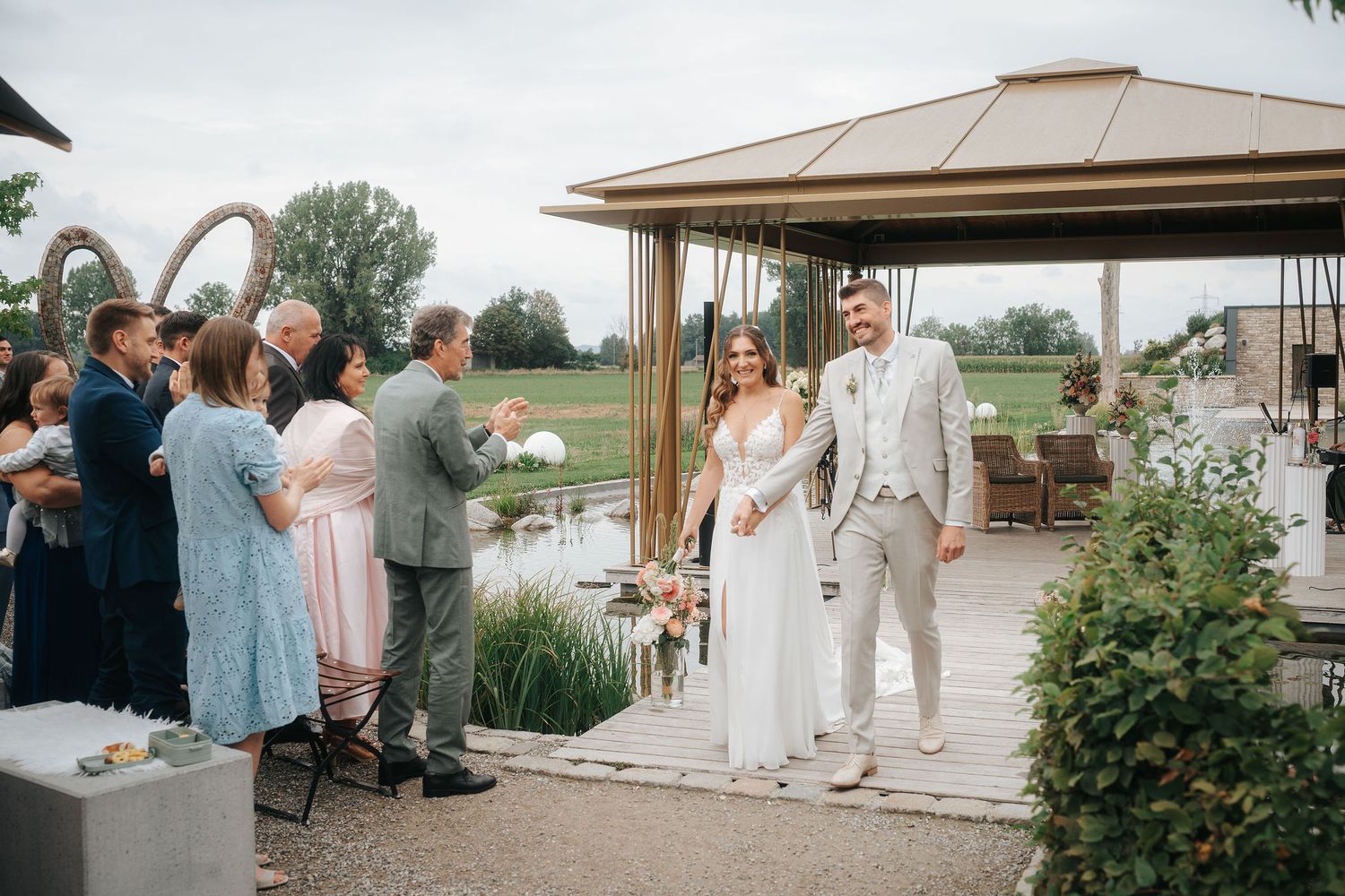 Bride in a white gown and groom in a light suit walk hand in hand toward guests at an outdoor garden wedding near a wooden gazebo.