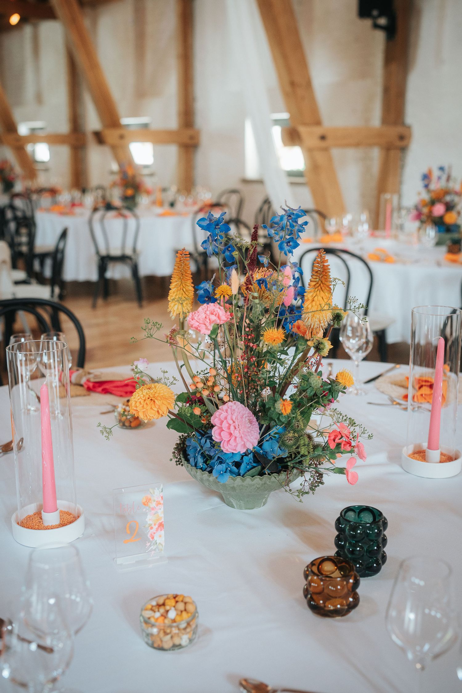Rustic wedding reception table with a colorful wildflower centerpiece, pink taper candles, and glassware in a timber-beamed barn venue.