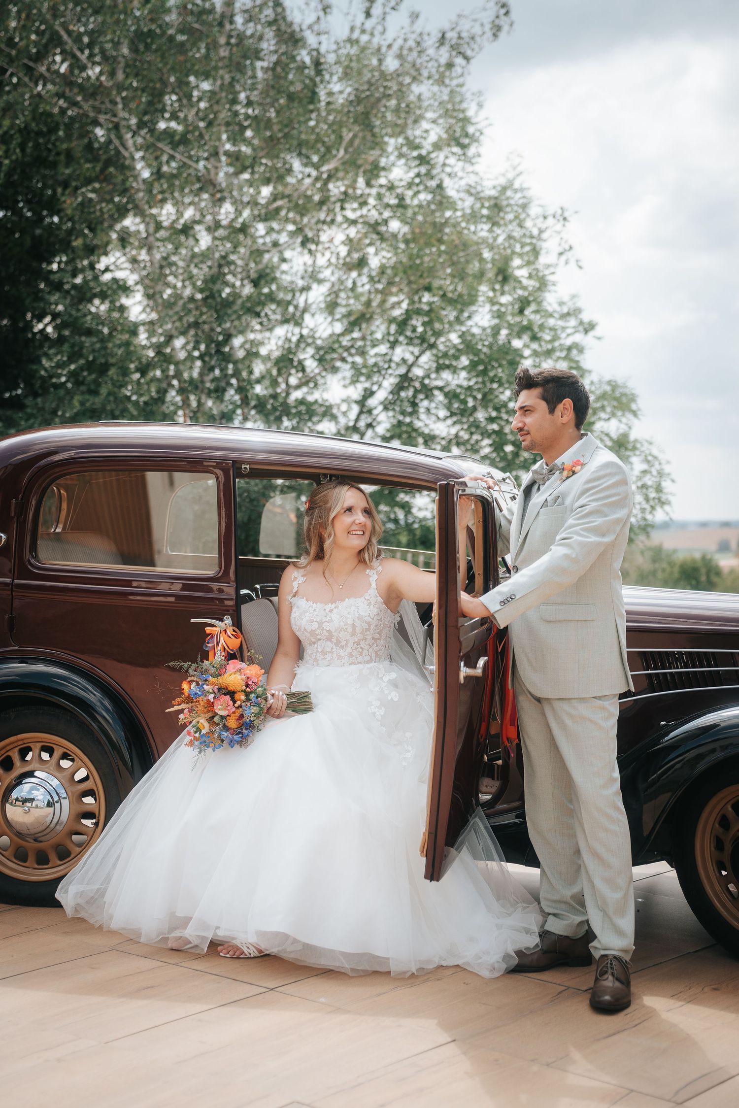 Bride in a white tulle wedding gown holding a colorful bouquet sits in a vintage car as the groom in a gray suit opens the door.