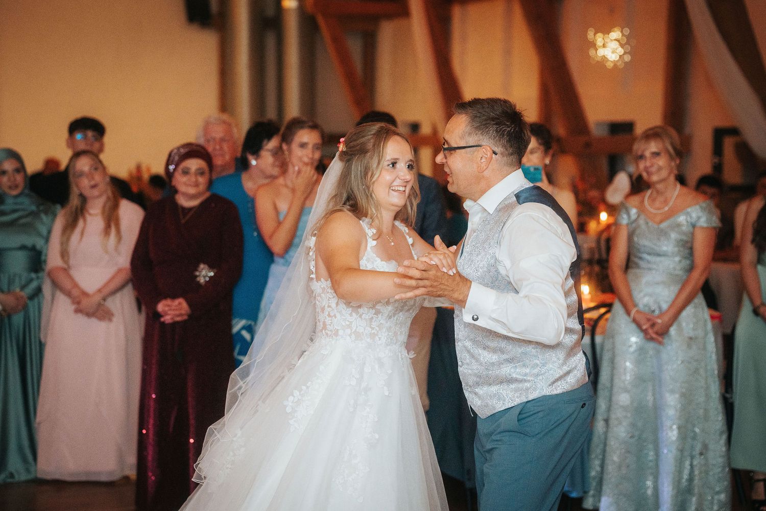 Bride in a white gown and groom in a vest share their first dance while smiling wedding guests in formal attire look on.
