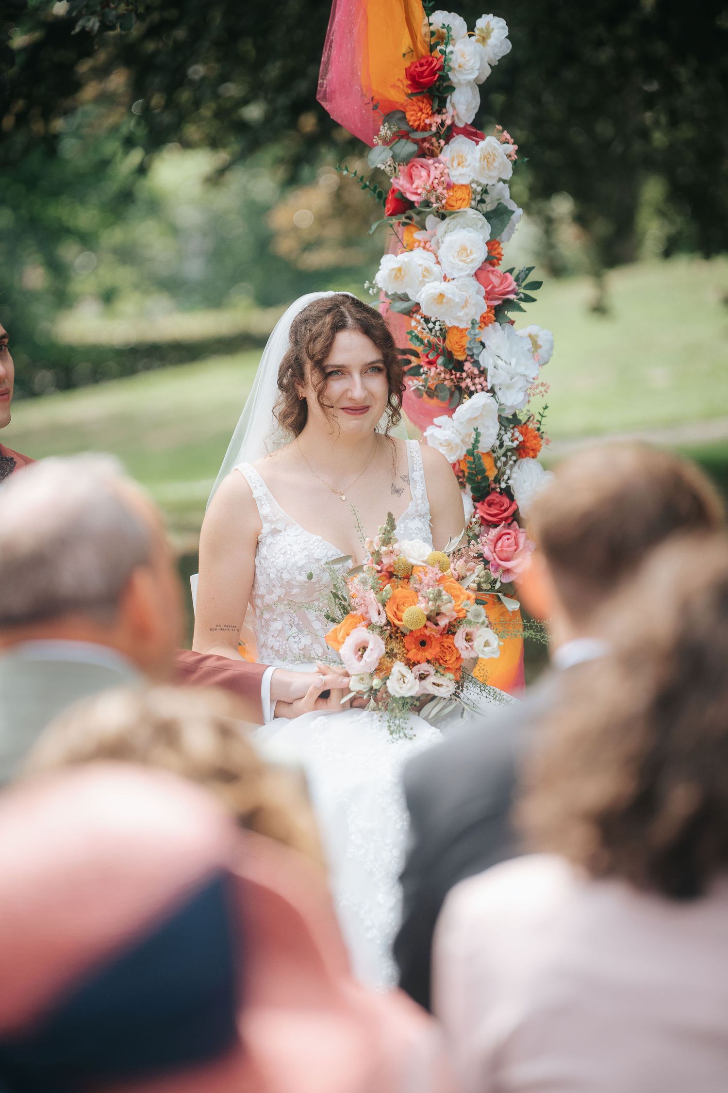 Smiling bride in a lace gown and veil holds an orange and pink bouquet beside a floral arch at an outdoor garden wedding ceremony.