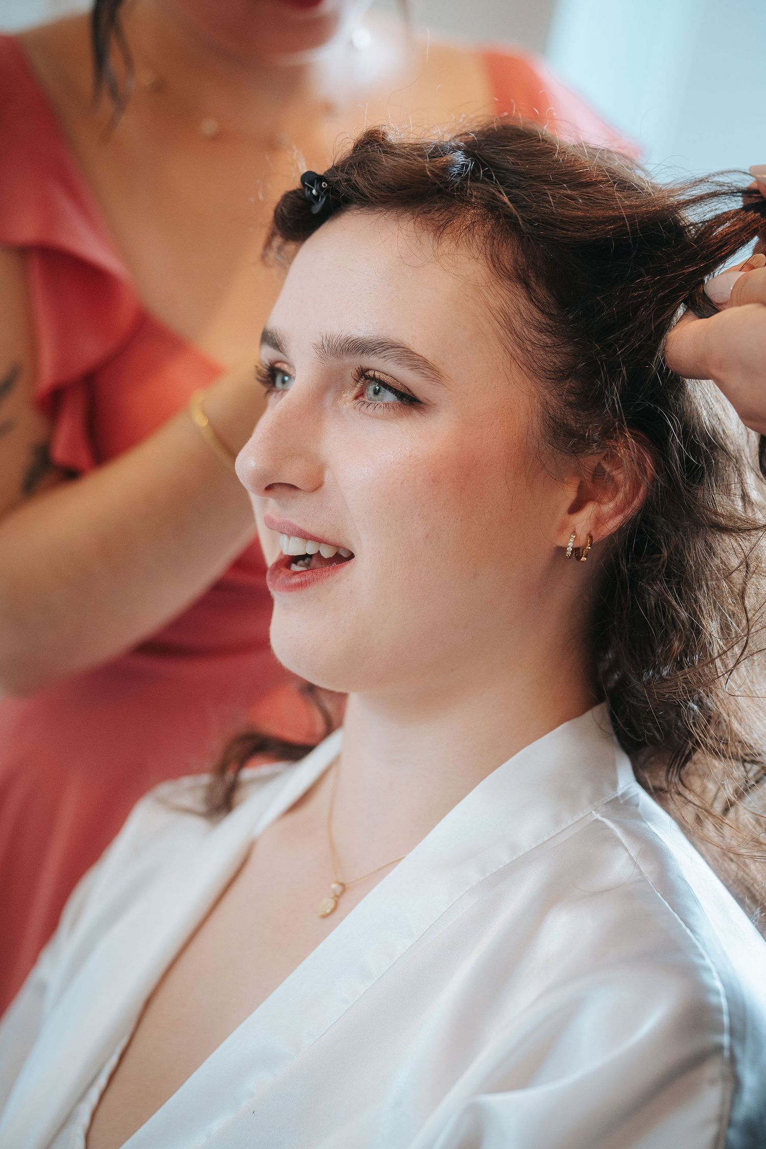 Smiling bride in a white satin robe has her wavy brown hair styled by a woman in a coral dress during wedding morning preparations.