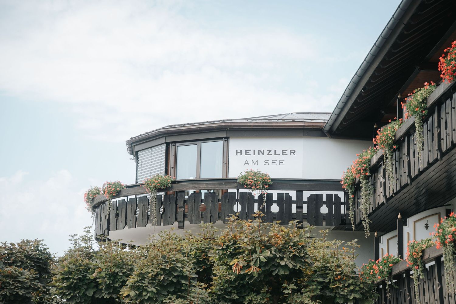 Rounded Heinzler am See hotel building with wooden balconies, flower boxes and a large window, surrounded by green trees under a blue sky.