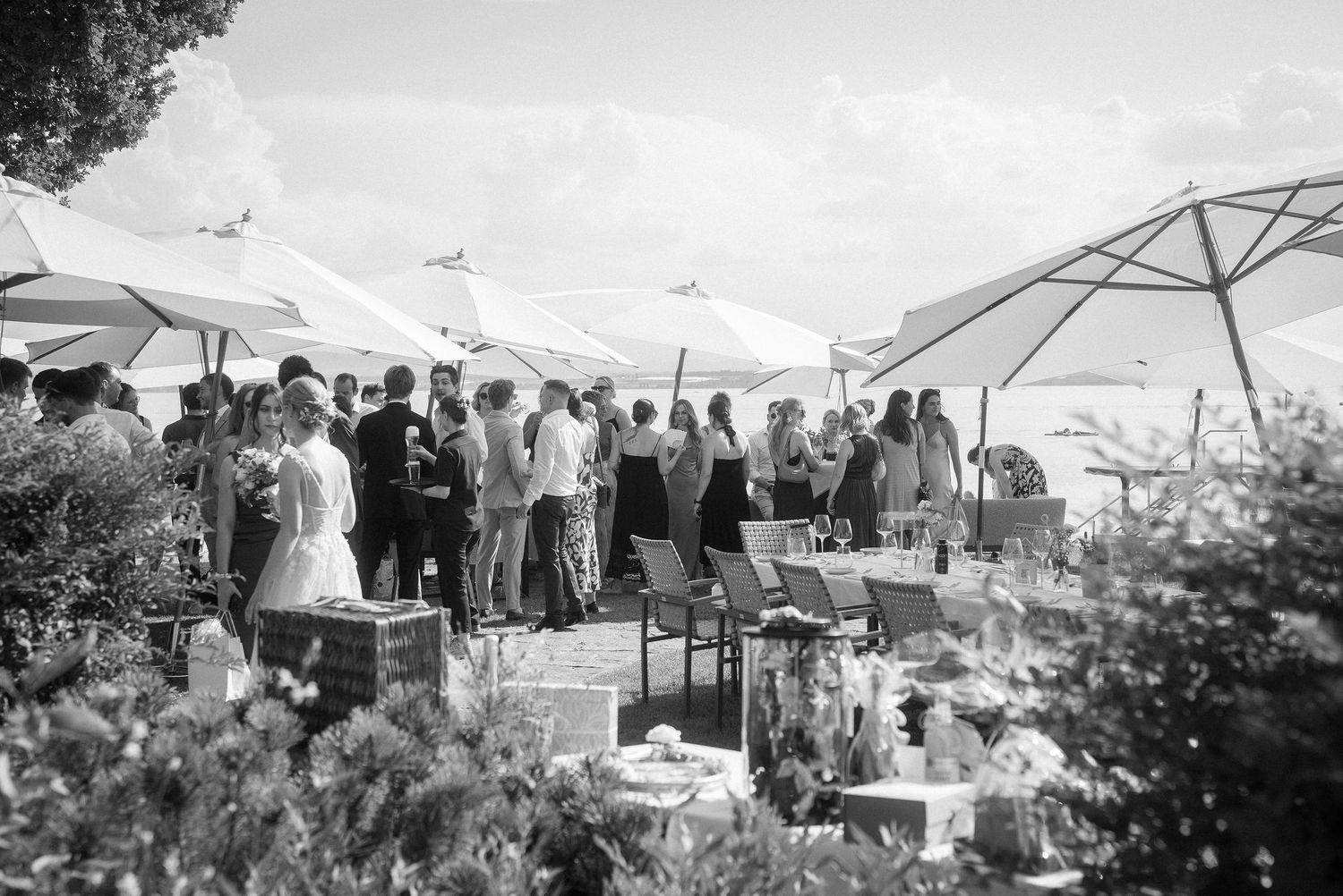 Black and white photo of wedding guests gathered outdoors beneath white patio umbrellas near a terrace with chairs and flower arrangements.