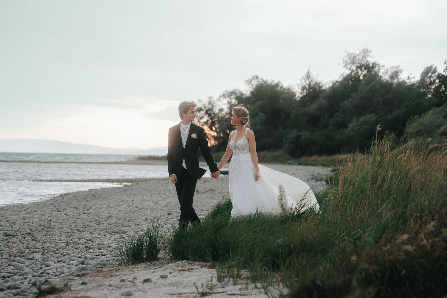 Bride in a white gown and groom in a dark suit hold hands while walking along a pebble beach with grassy dunes at sunset.