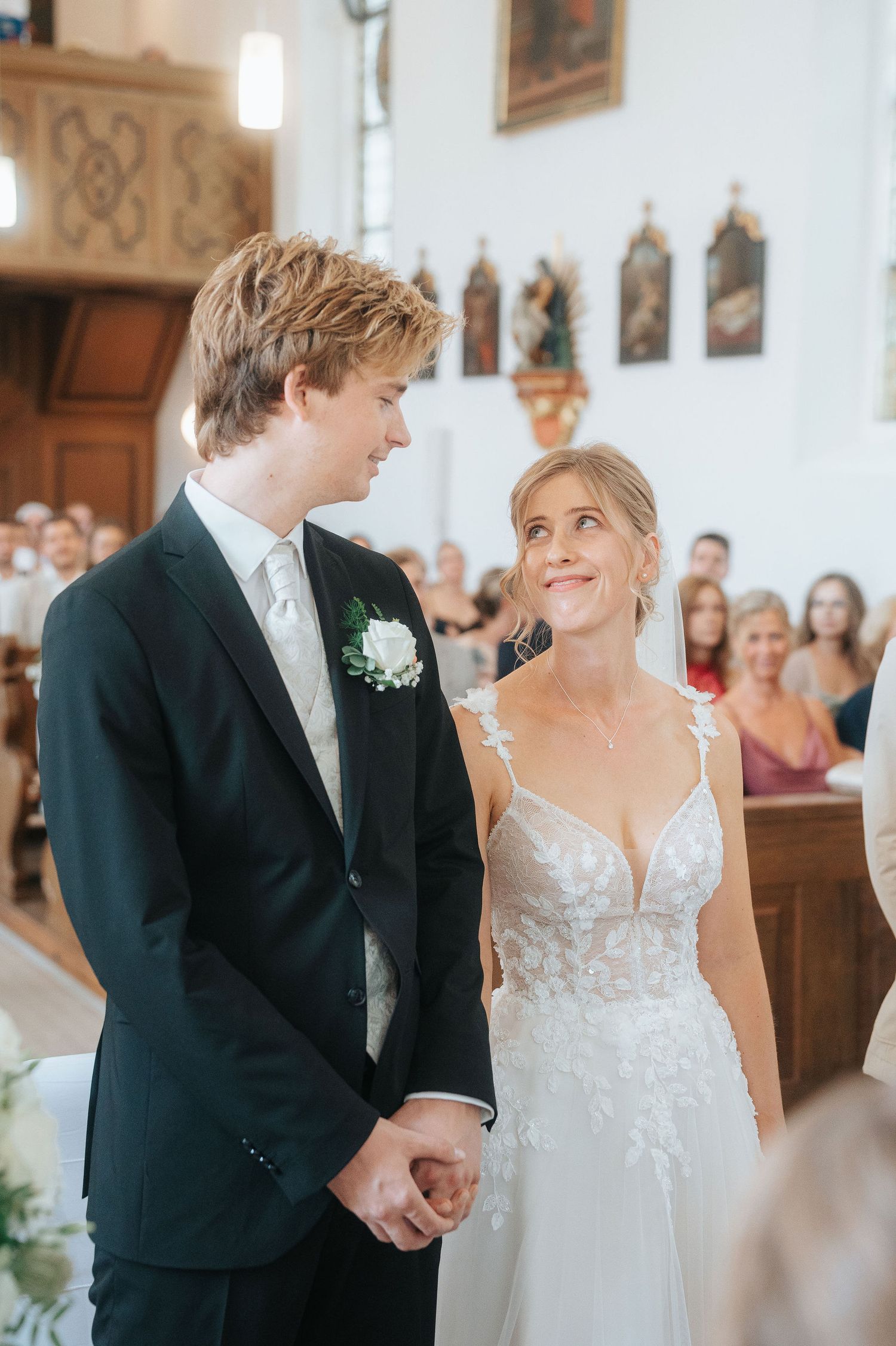 Bride in a lace wedding gown and groom in a dark suit with white boutonniere hold hands during a church ceremony as guests look on.