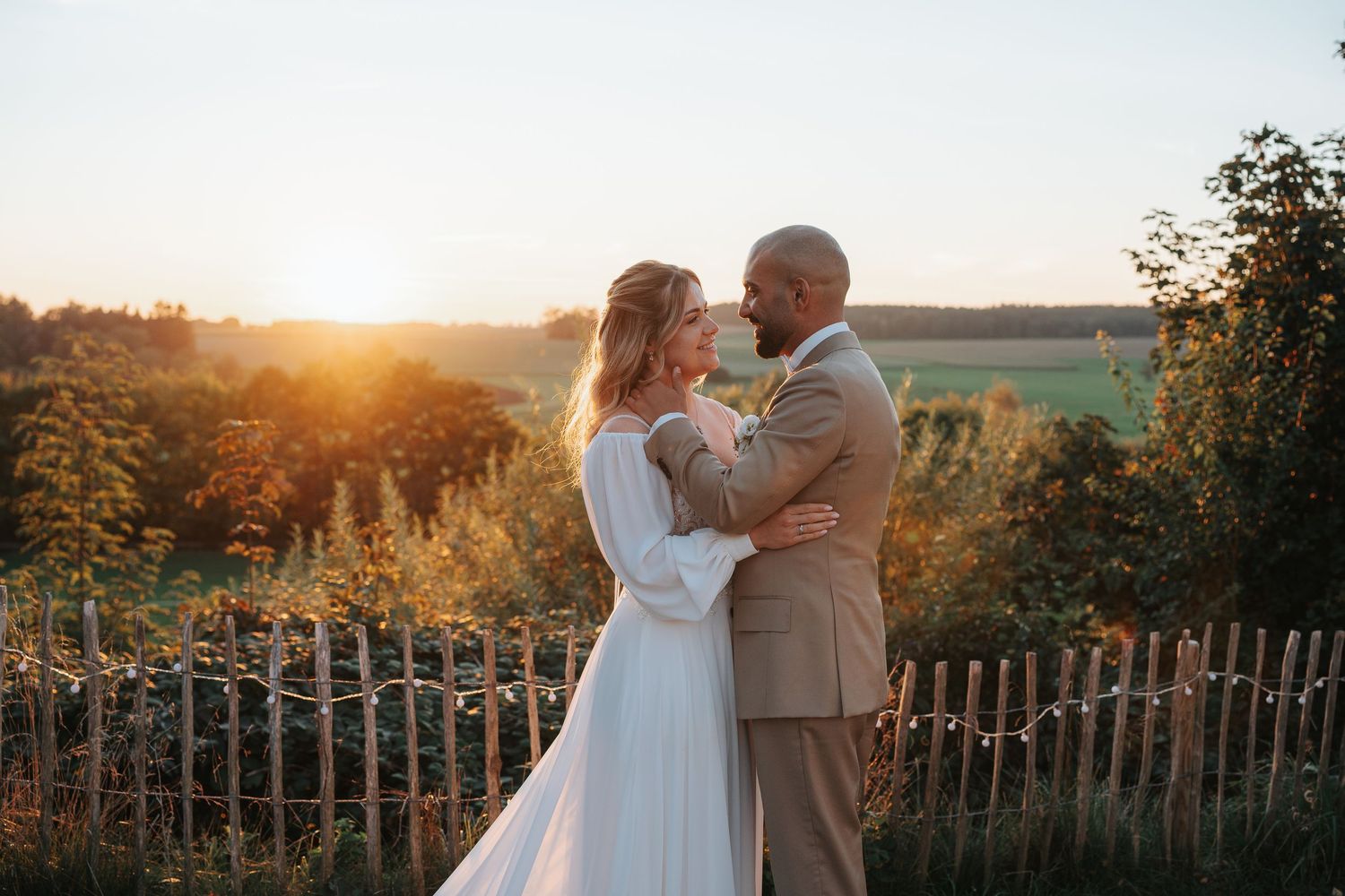 A couple shares a romantic moment at sunset with a rustic wooden fence and golden sunlight in the background.