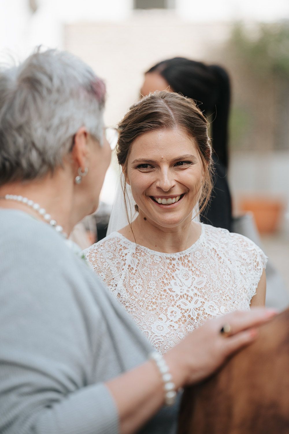 A radiant bride in a white lace dress sharing a joyful moment with a guest wearing pearls at an outdoor celebration.