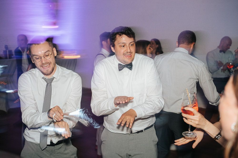 Wedding guests dance and laugh together on a dance floor illuminated by purple lighting at an evening reception.