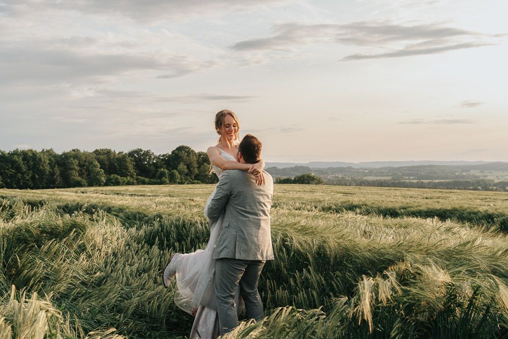 Loving couple sharing a tender moment while standing in a hilltop wheat field with scenic countryside views.
