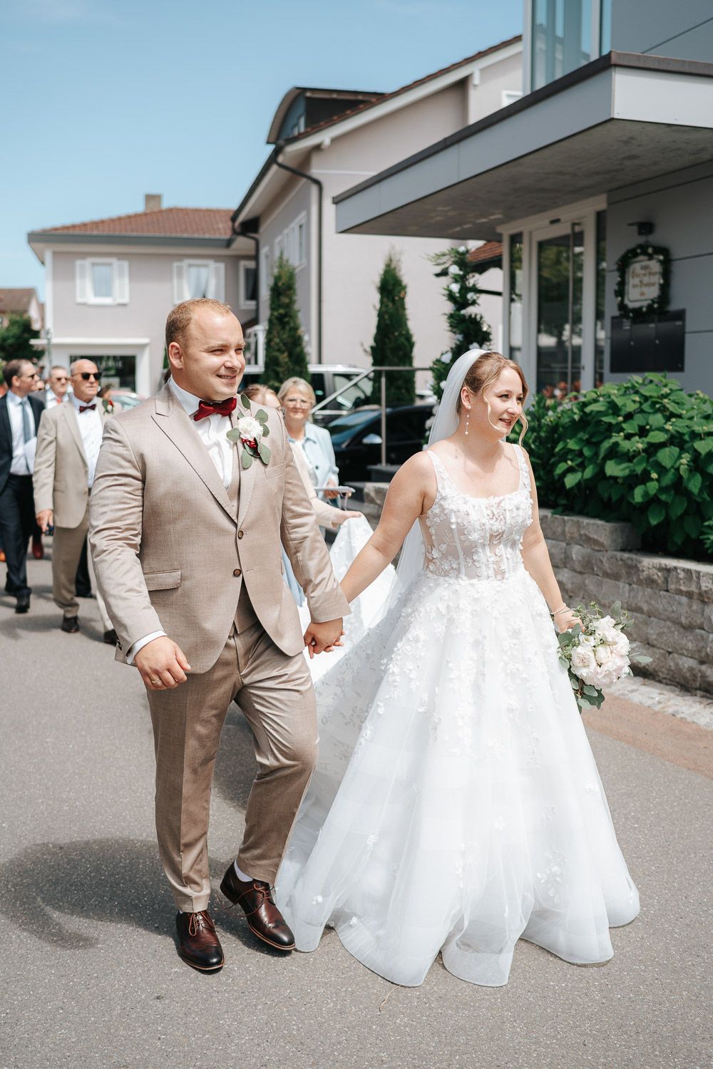 A couple celebrates their wedding day, with the bride in a white gown and groom in tan suit walking together outdoors.