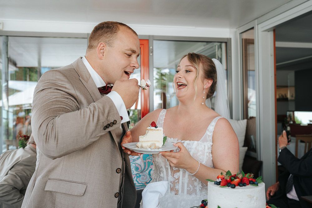 A newlywed couple shares a joyful moment while eating wedding cake together during their reception celebration.