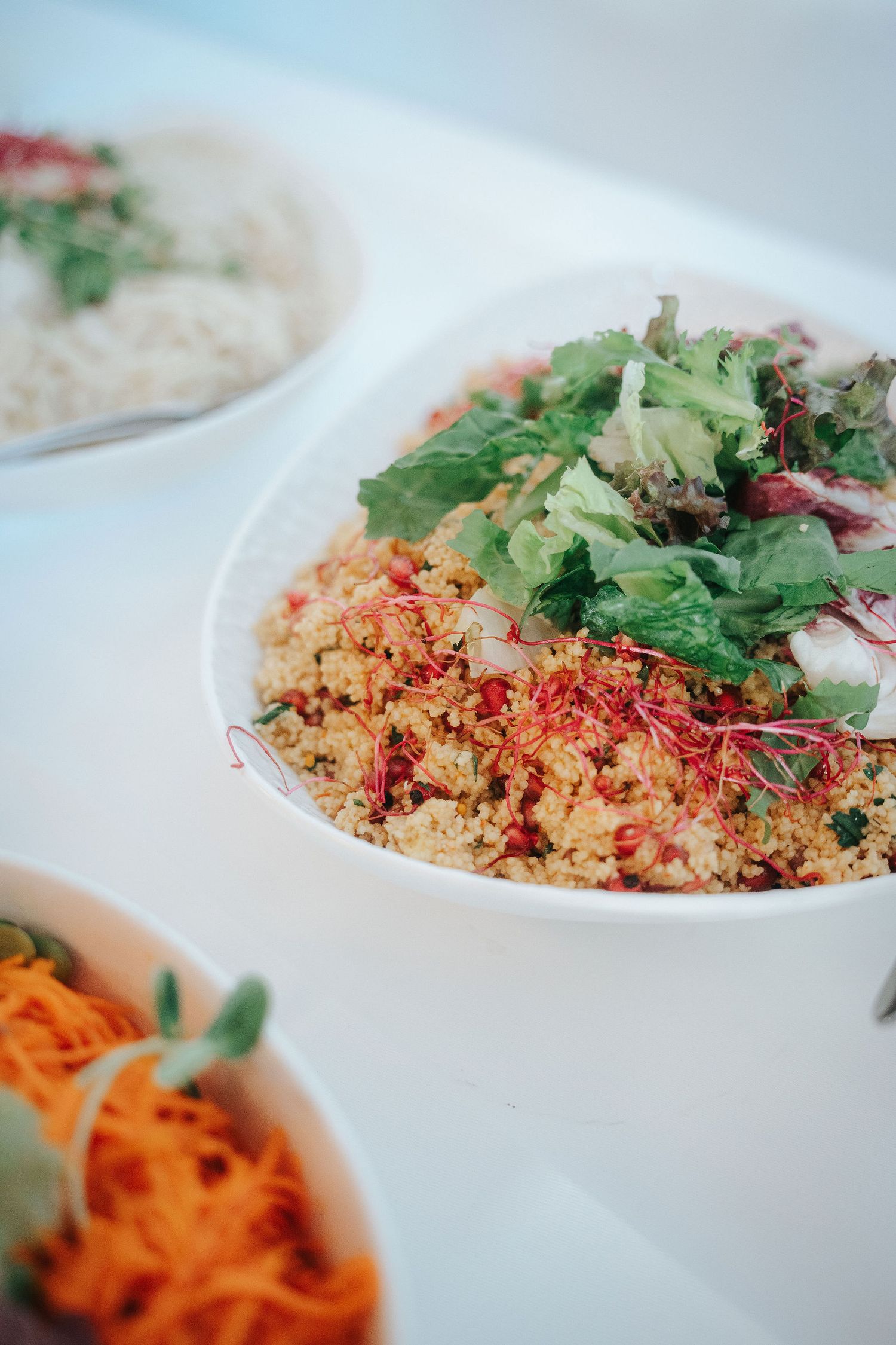 Fresh grain salad topped with leafy greens and red sprouts in a white bowl, served beside a carrot salad and a rice dish.