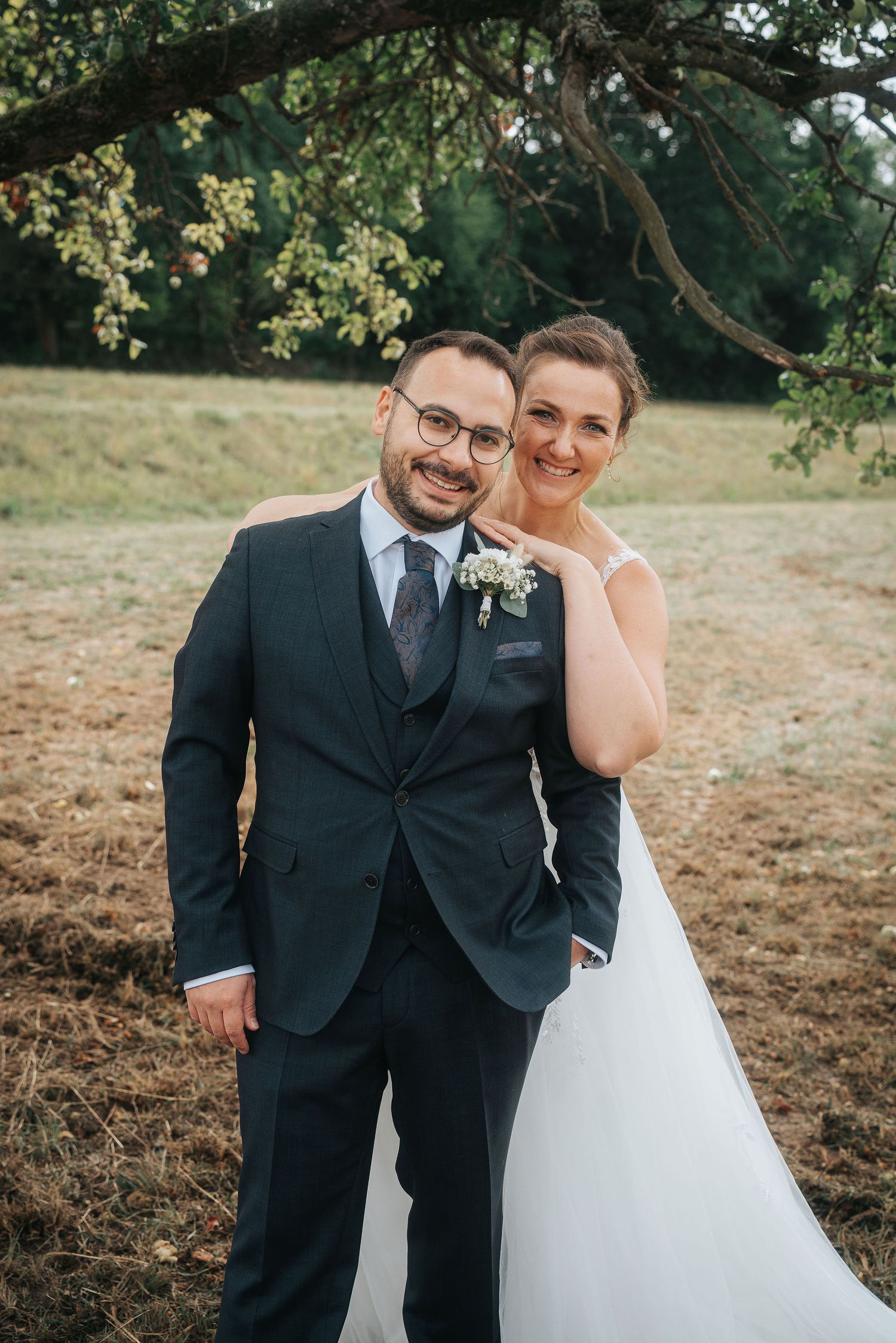 Smiling bride in a white wedding gown embraces her groom in a navy three-piece suit with a floral boutonniere in a sunny countryside field.
