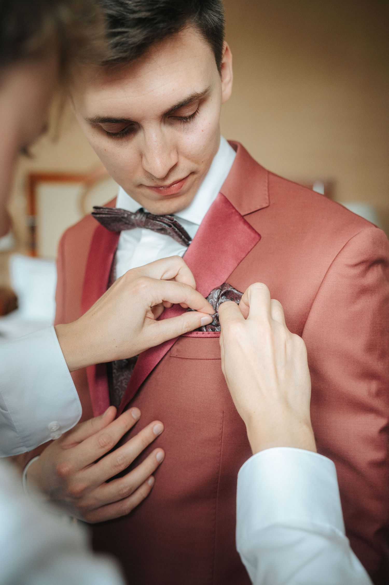 Groomsman in a coral pink suit and brown bow tie has a boutonniere pinned to his lapel by two helping hands before the wedding.
