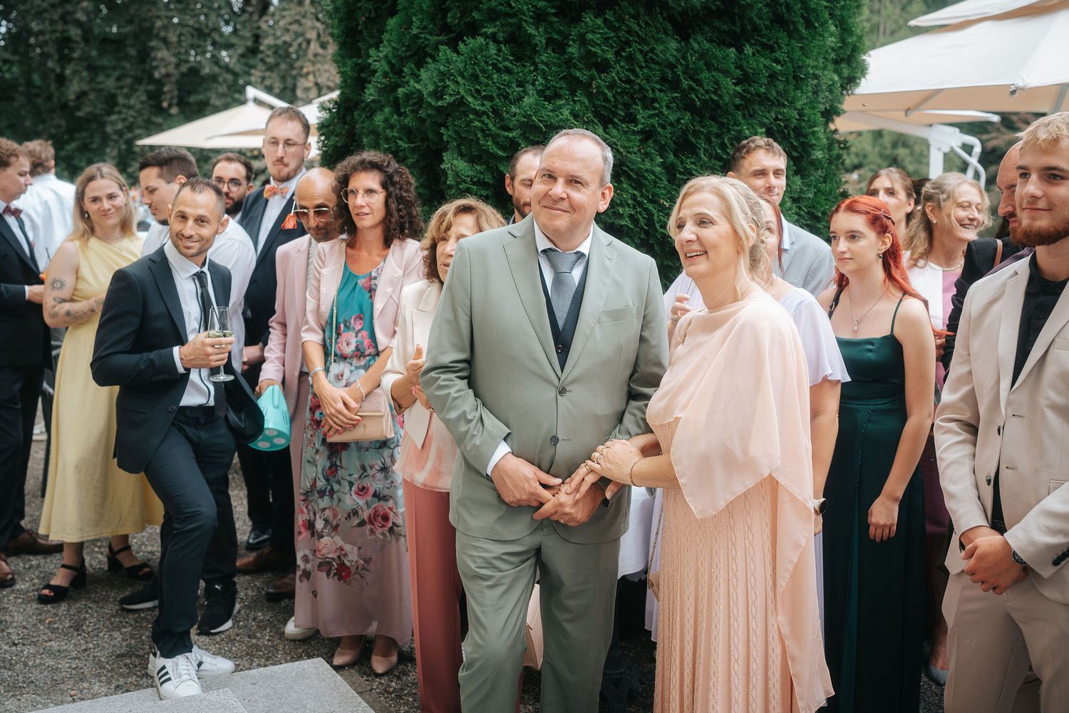 Smiling wedding guests in elegant attire gather outdoors, with a man in a gray suit and a woman in a peach dress standing at the front.