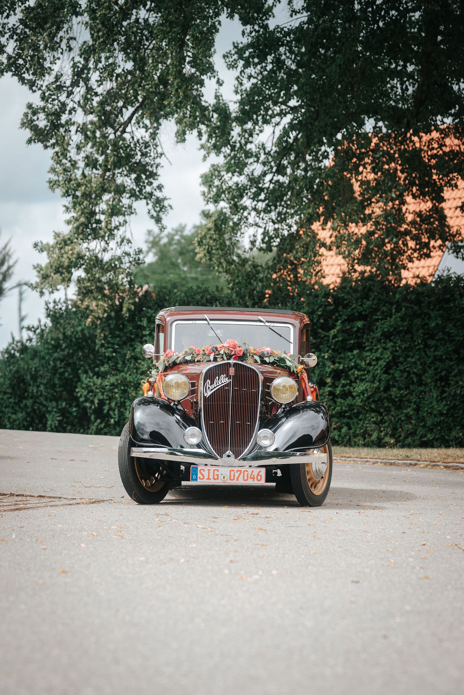 Vintage burgundy wedding car decorated with a floral garland parked on a quiet road beneath leafy green trees.