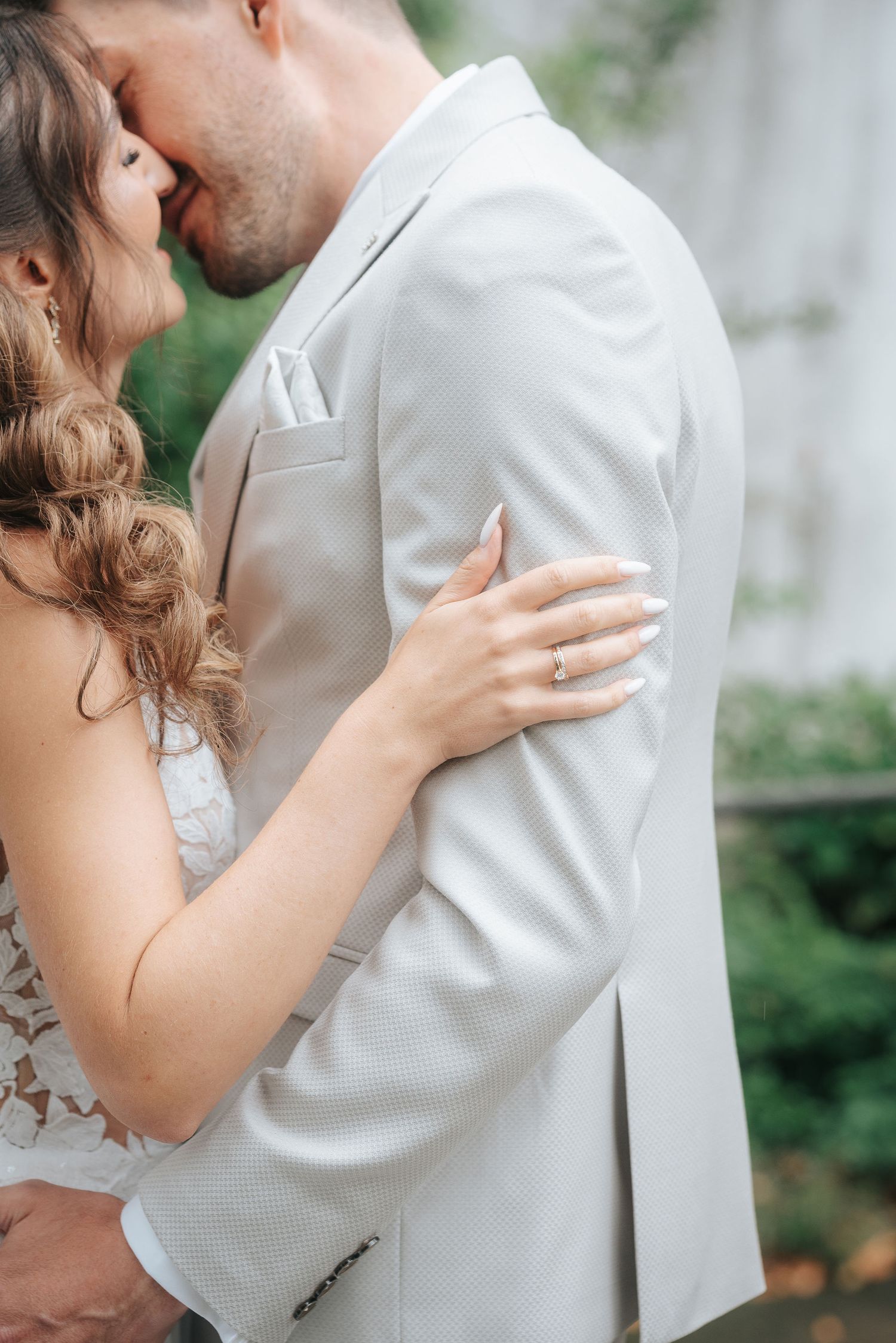 Bride in a lace wedding dress embraces a groom in a light beige suit, her ring-adorned hand on his shoulder, in a green garden.