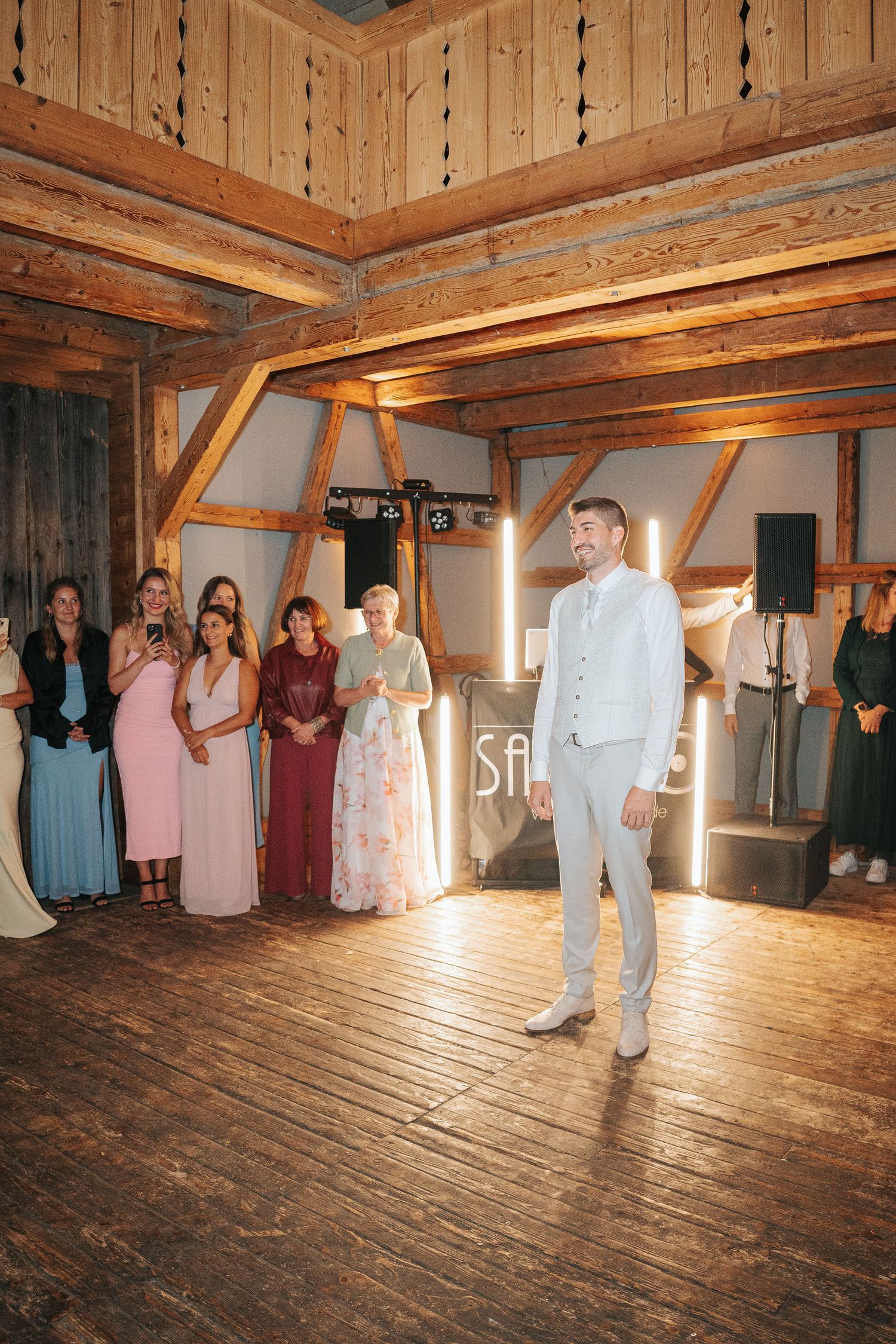 A groom in a light vest and trousers stands on a wooden dance floor as wedding guests watch inside a rustic timber barn venue.