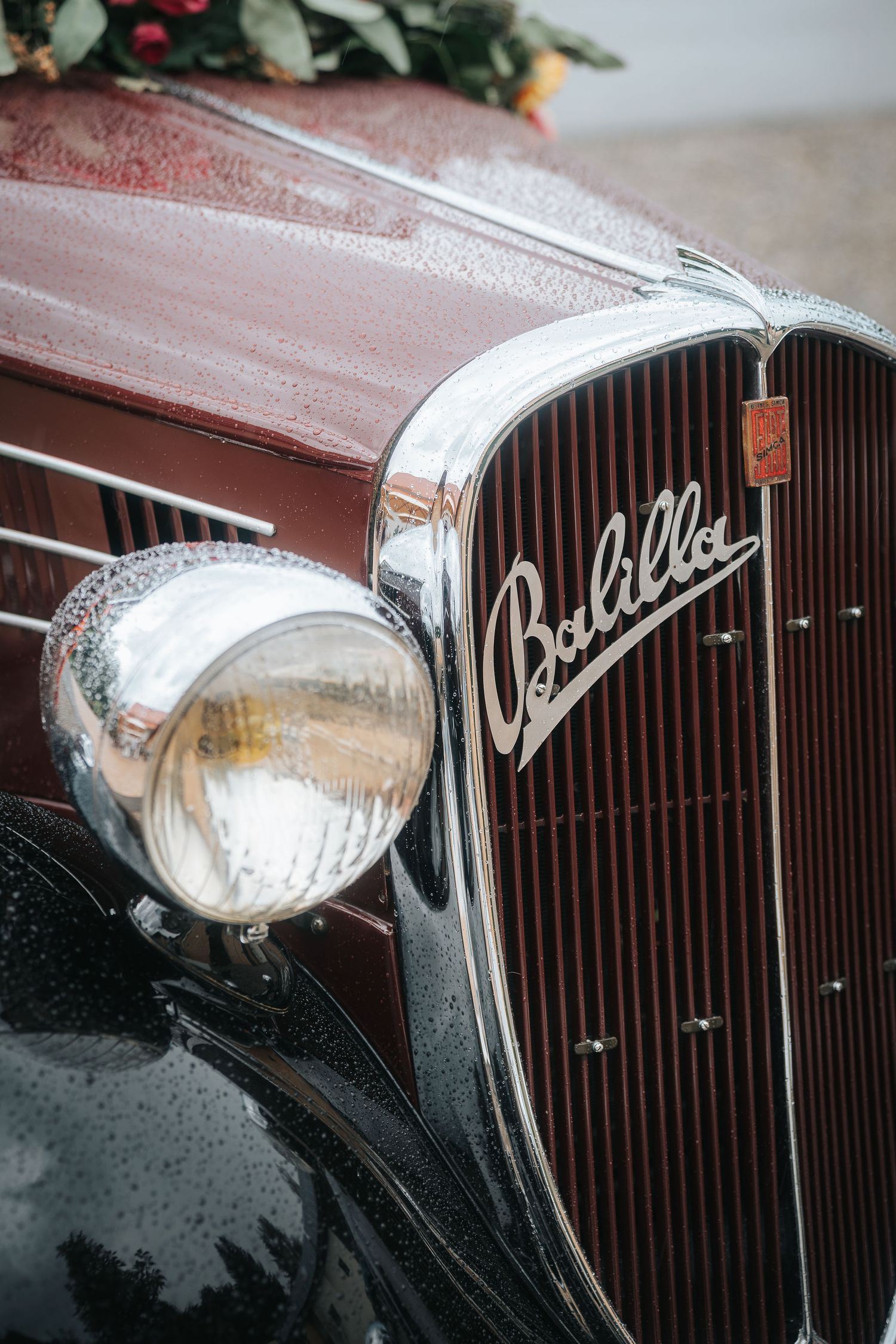 Close-up of a vintage burgundy Fiat Balilla car with chrome headlight, vertical grille and script badge, dotted with raindrops.