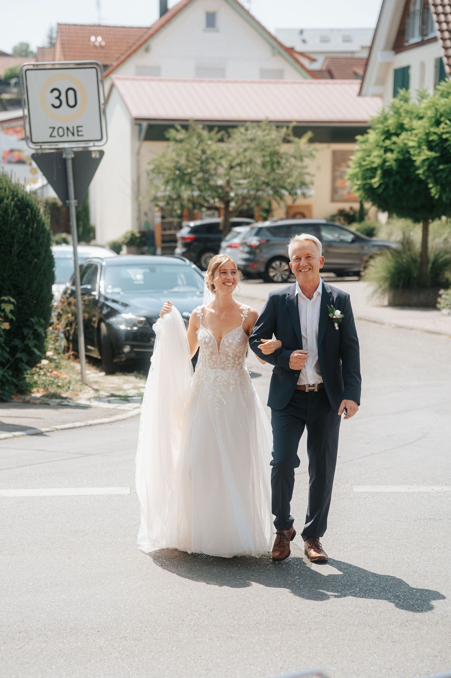 Bride in a lace wedding gown walks arm in arm with a man in a navy suit down a sunny residential street past a 30 zone sign.