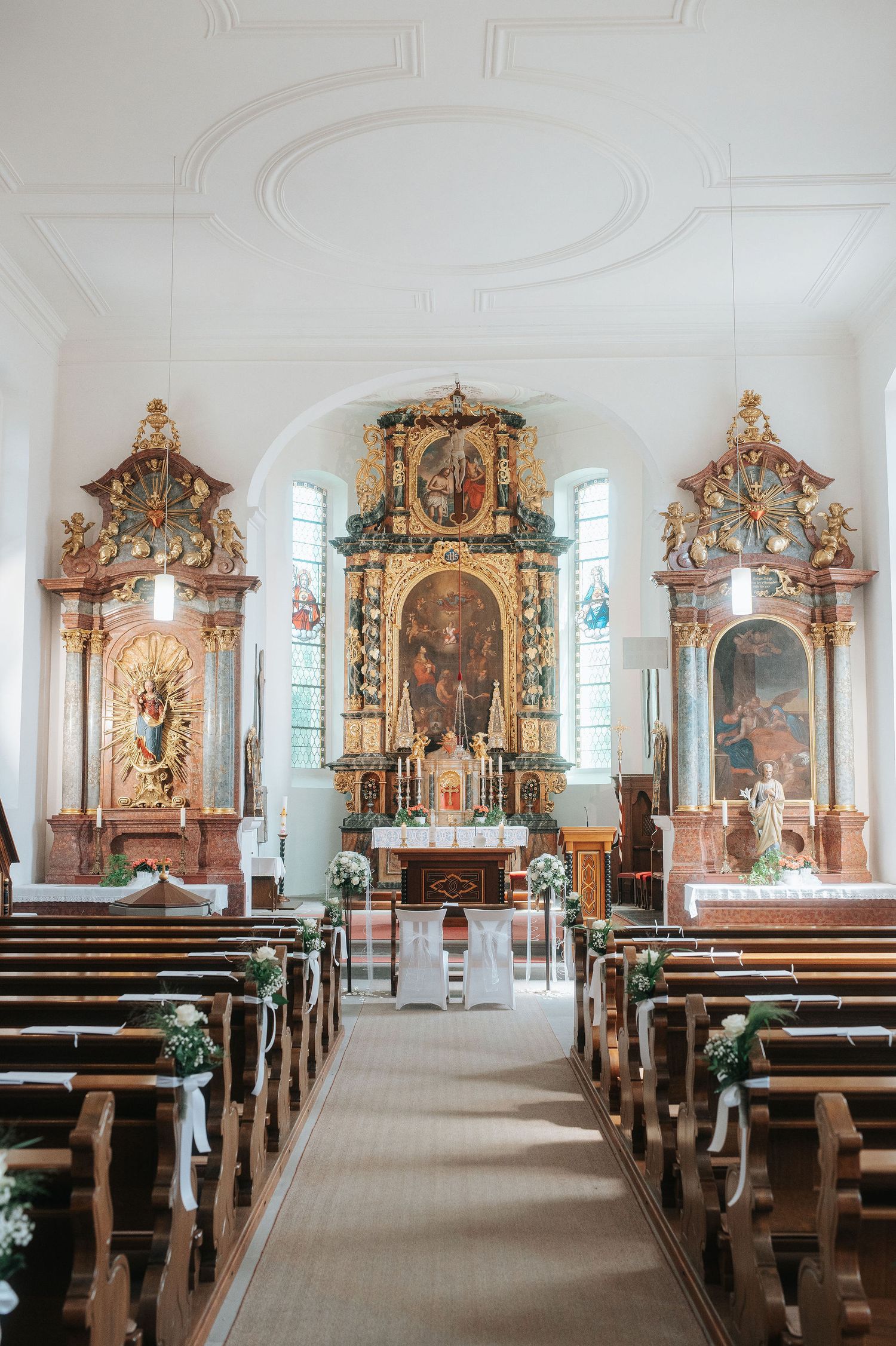 Ornate Baroque church interior decorated with flowers and white ribbons on wooden pews, leading to a golden altar and side altars.
