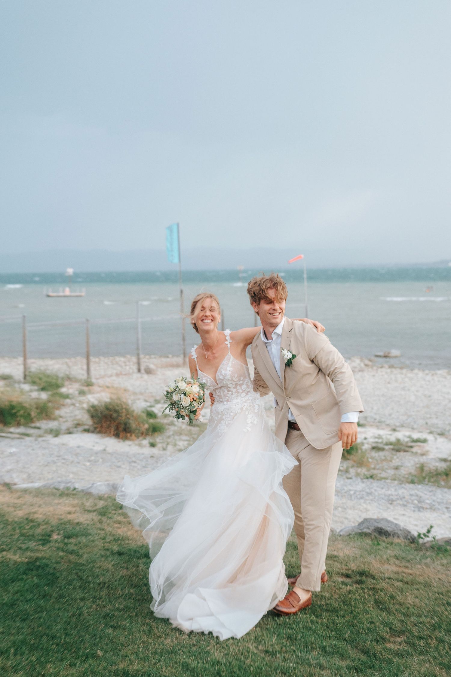 Smiling bride in a flowing tulle gown and groom in a tan suit embrace on a grassy lakeside shore on their wedding day.