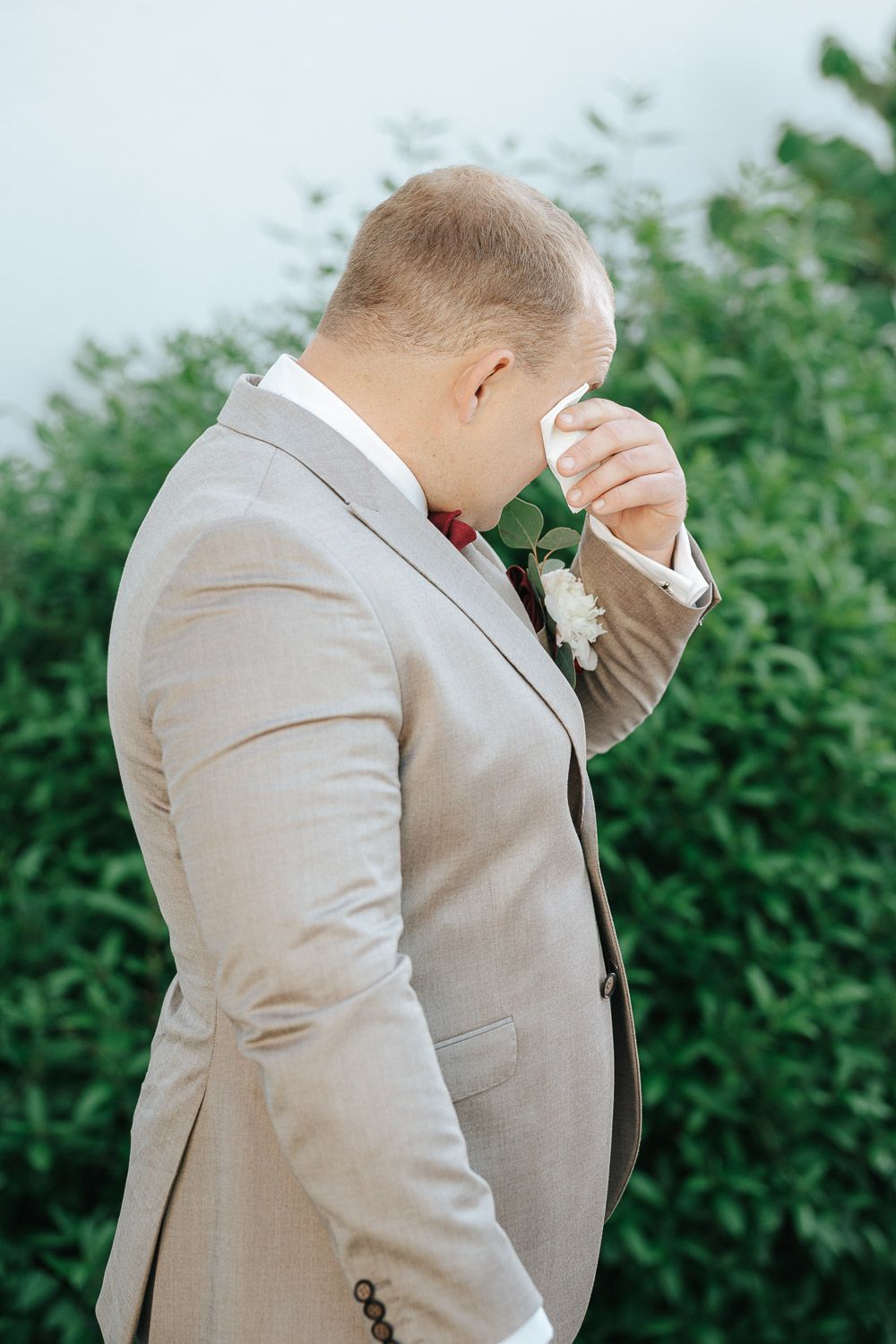 Emotional wedding moment captured as person in light beige suit wipes tears away against green foliage background.