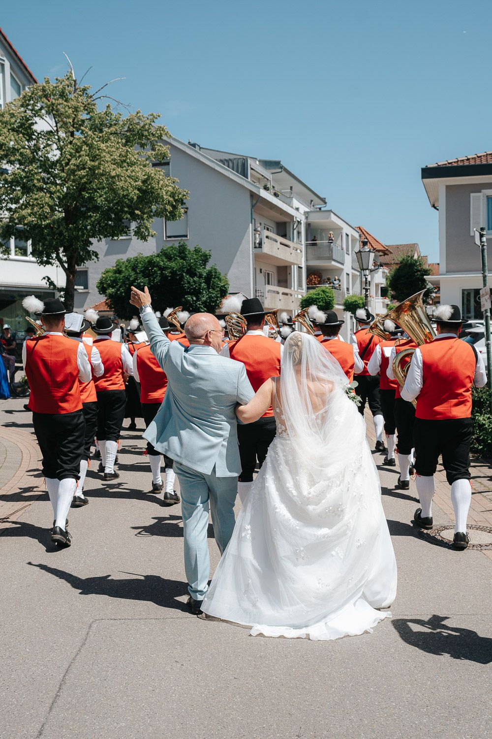Traditional marching band in red uniforms lead a newlywed couple down a residential street during a wedding celebration.