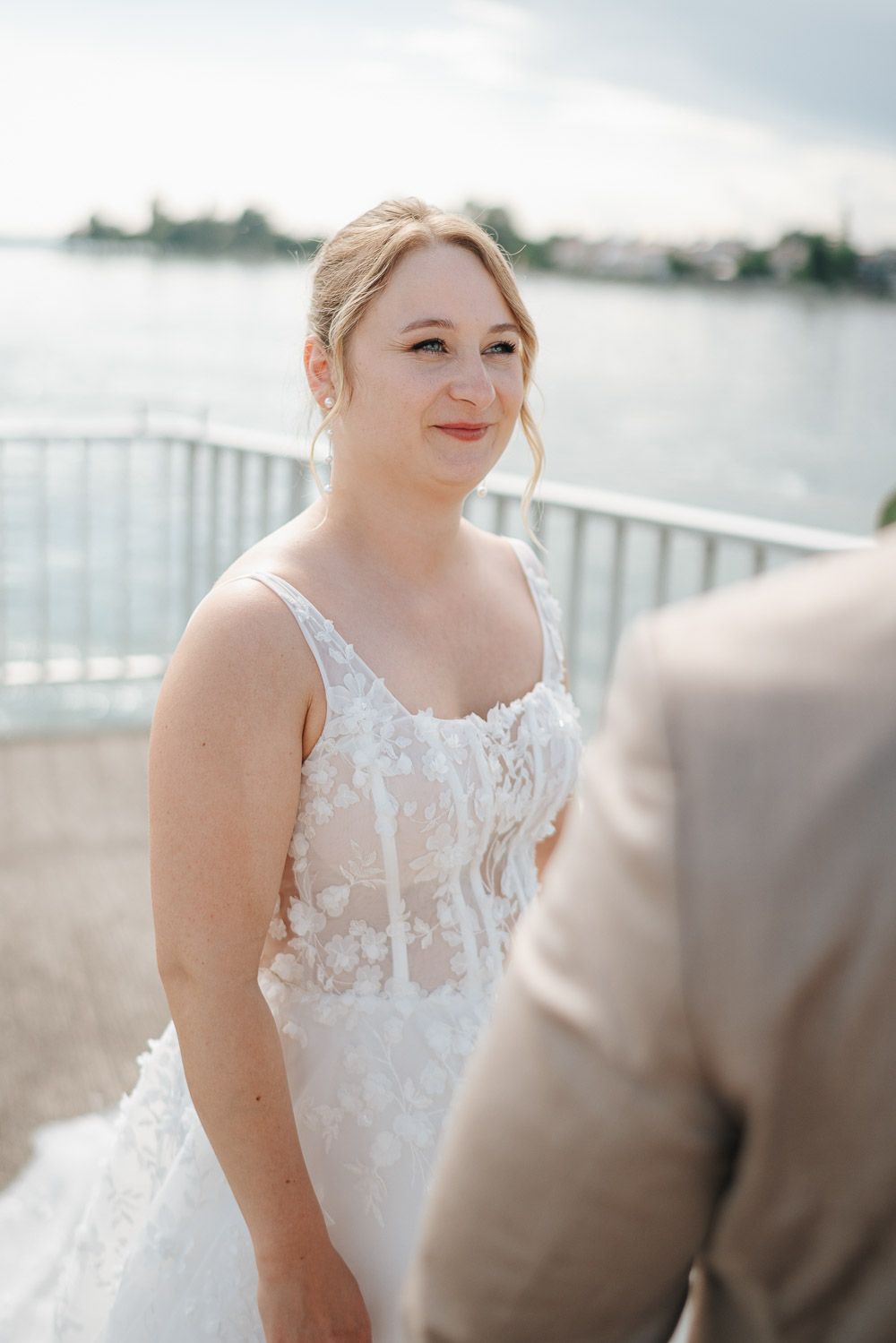 Beautiful wedding dress with delicate lace details and sheer overlay shown in outdoor waterfront setting.