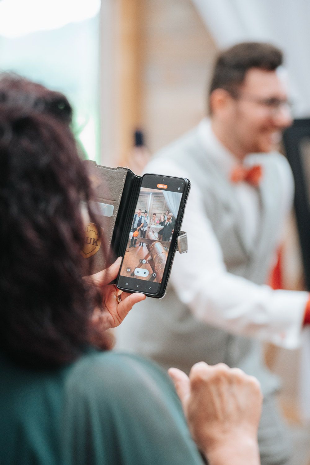 Someone taking a photo on their smartphone at a gathering as a person in a white shirt and red tie smiles in the background.