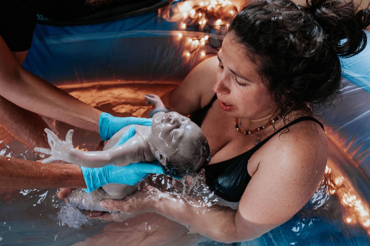 Woman receives therapeutic water birth care with soft lights and blue water in birthing pool.