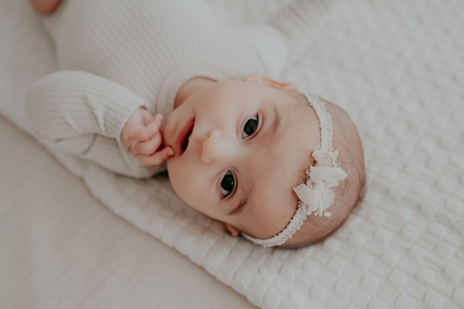 Close-up of an infant wearing a delicate white headband while lying on soft bedding.