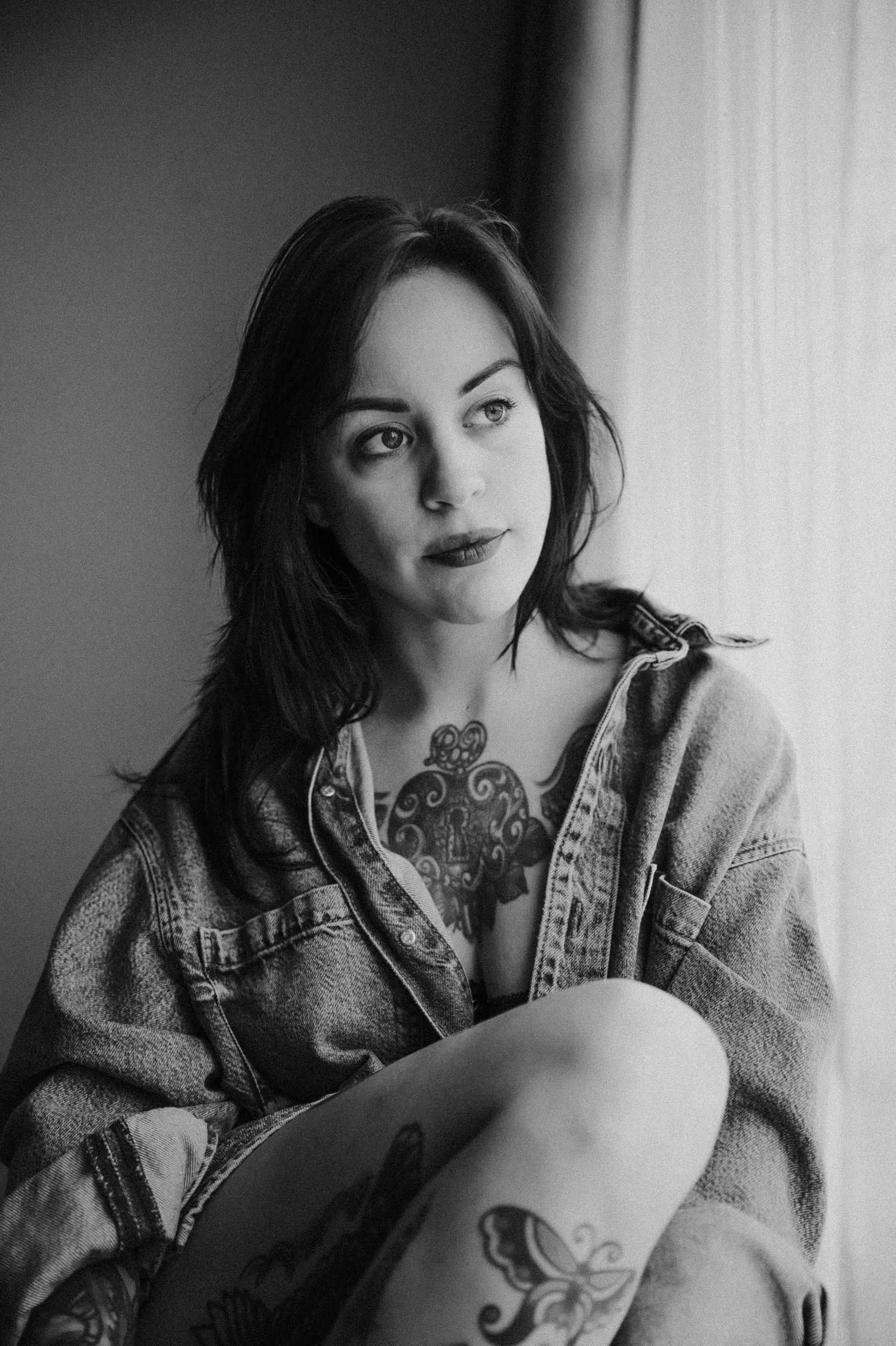 Black and white portrait of a tattooed woman in denim jacket sitting by a window with soft natural light.
