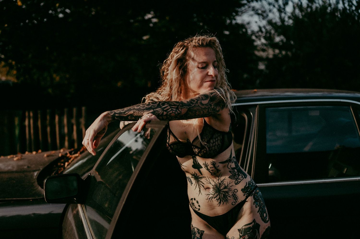 A person in dark clothing poses with attitude next to a vintage car during golden hour.