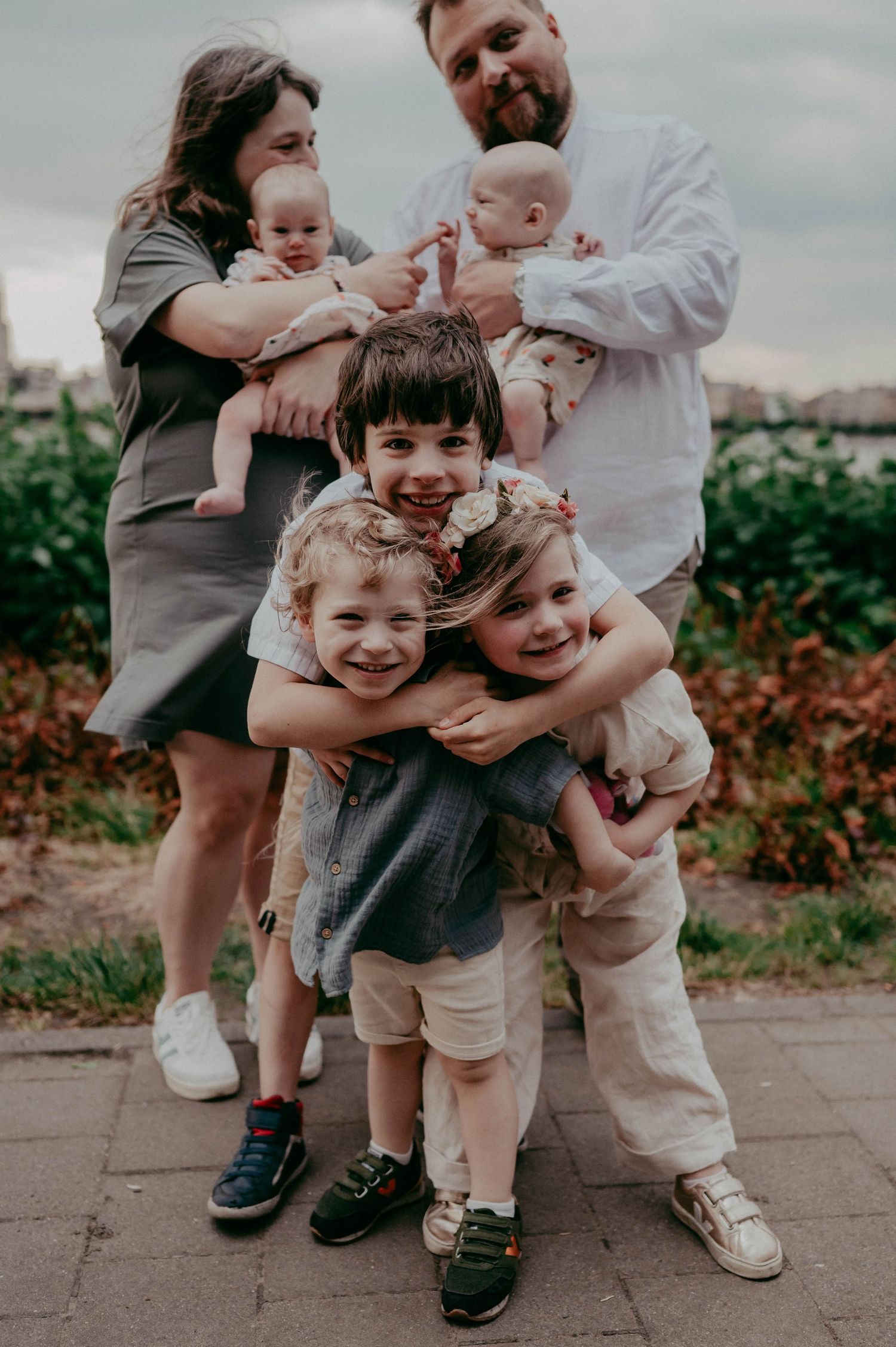 Playful outdoor family portrait with children hugging and twins in casual attire.