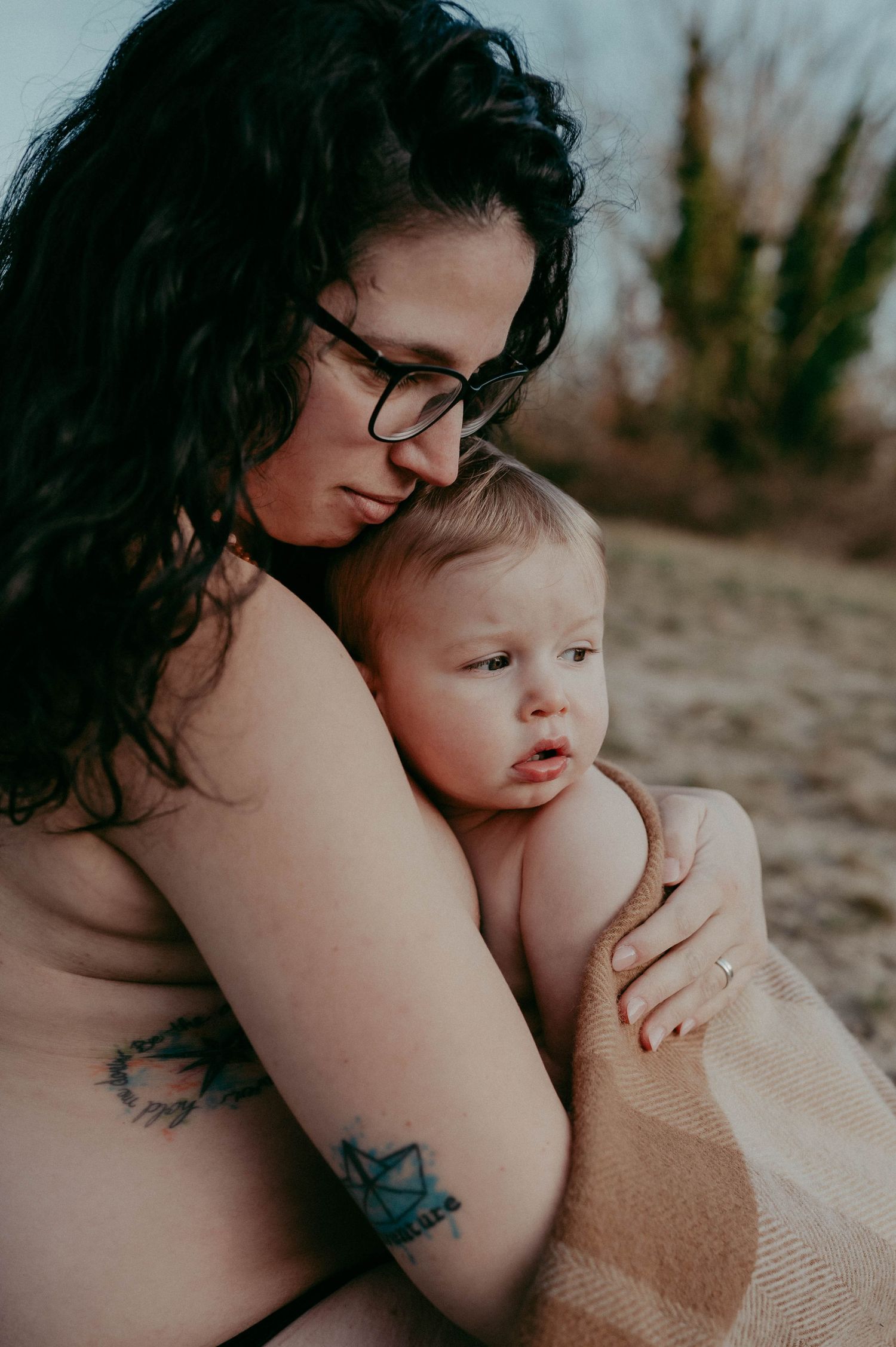 Artistic portrait showing intimate moment between parent with tattoos and baby against natural backdrop.