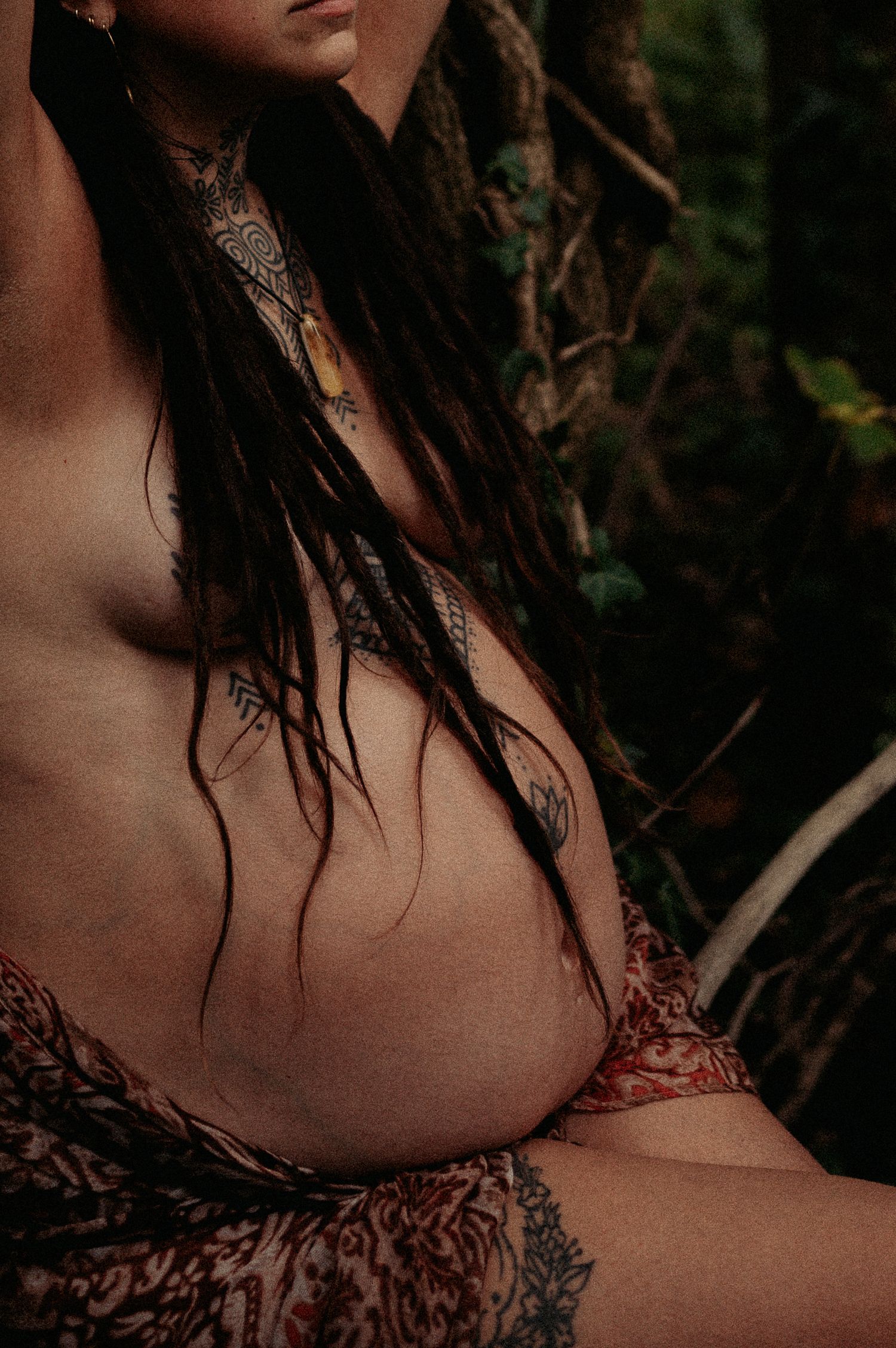 Artistic close-up maternity portrait showing tattoos and dreadlocks in natural setting.