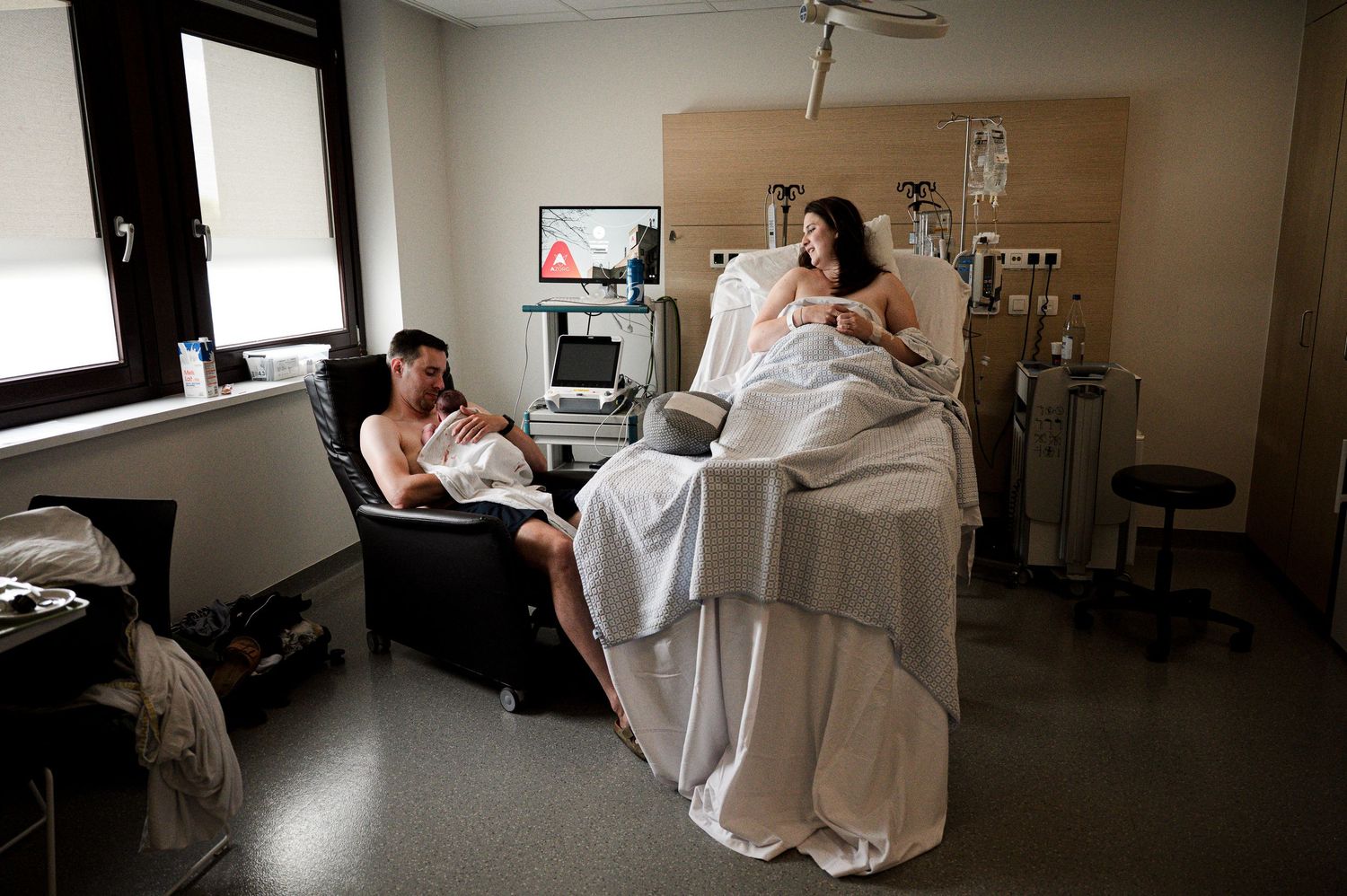 A patient rests in a hospital room bed while a visitor sits in a chair nearby with medical equipment in the background.
