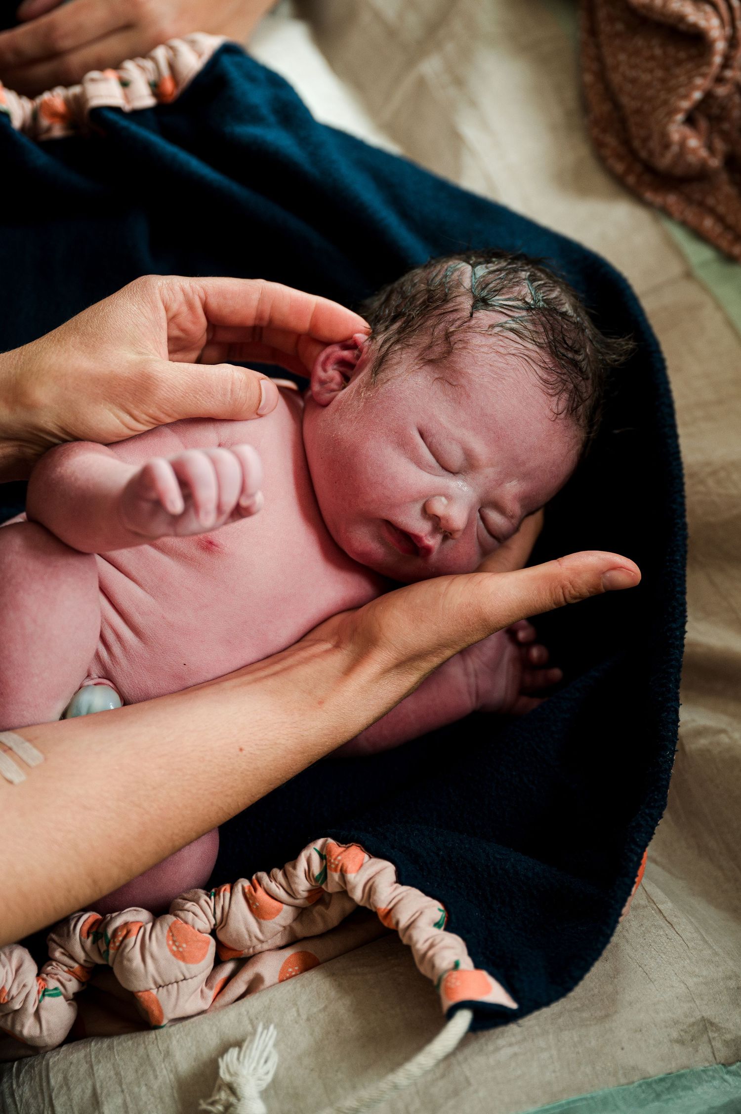 A newborn baby rests on gentle hands against a dark blue blanket with patterned fabric.