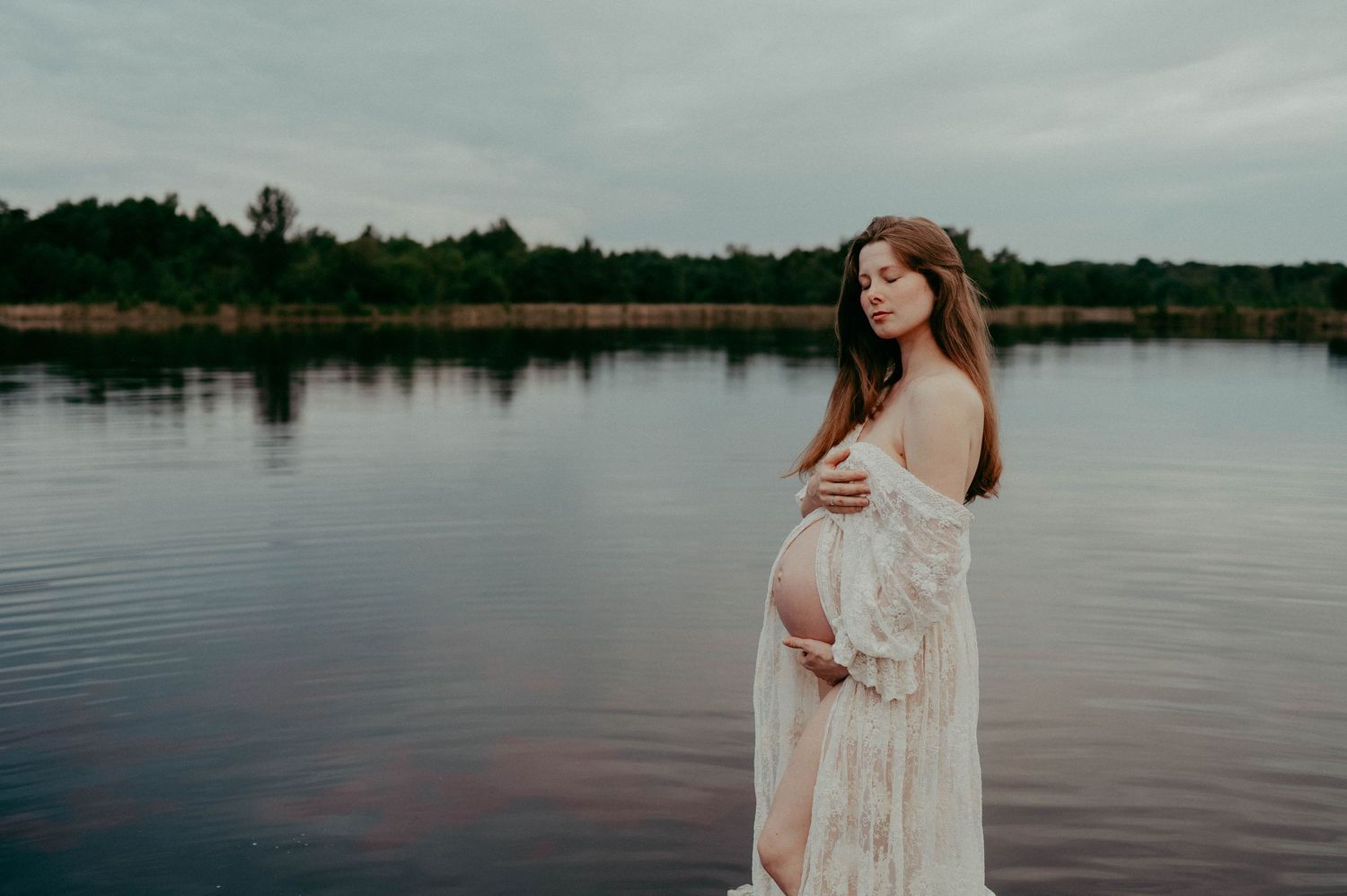 Ethereal maternity photo of an expectant mother in a white lace dress standing by a serene lake at dusk.