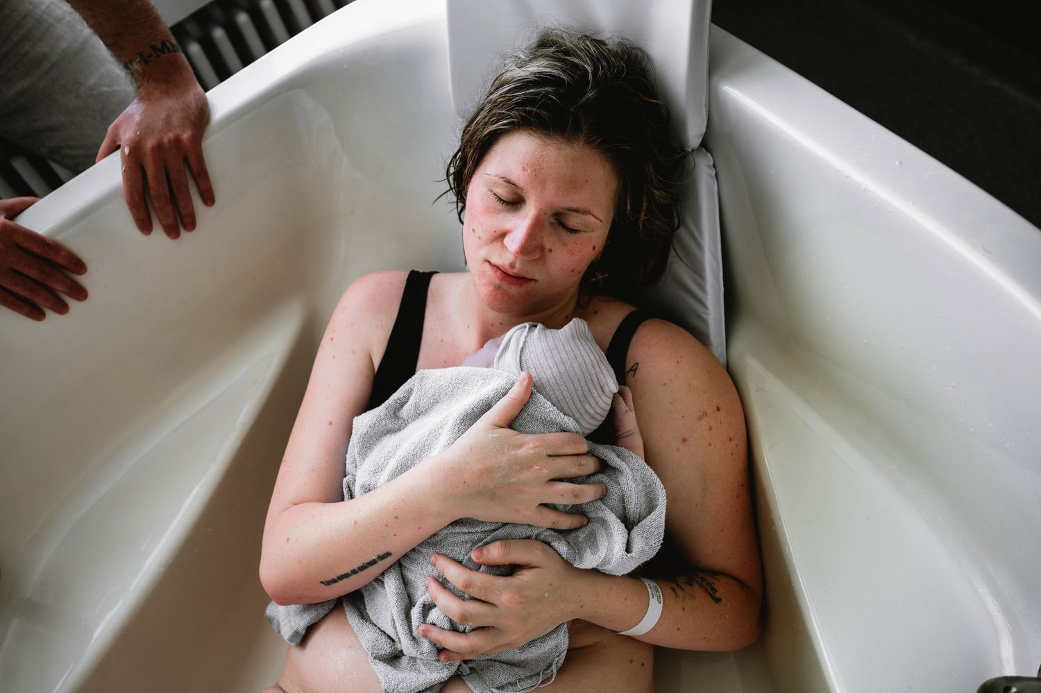 New parent rests in bathtub while holding swaddled newborn.