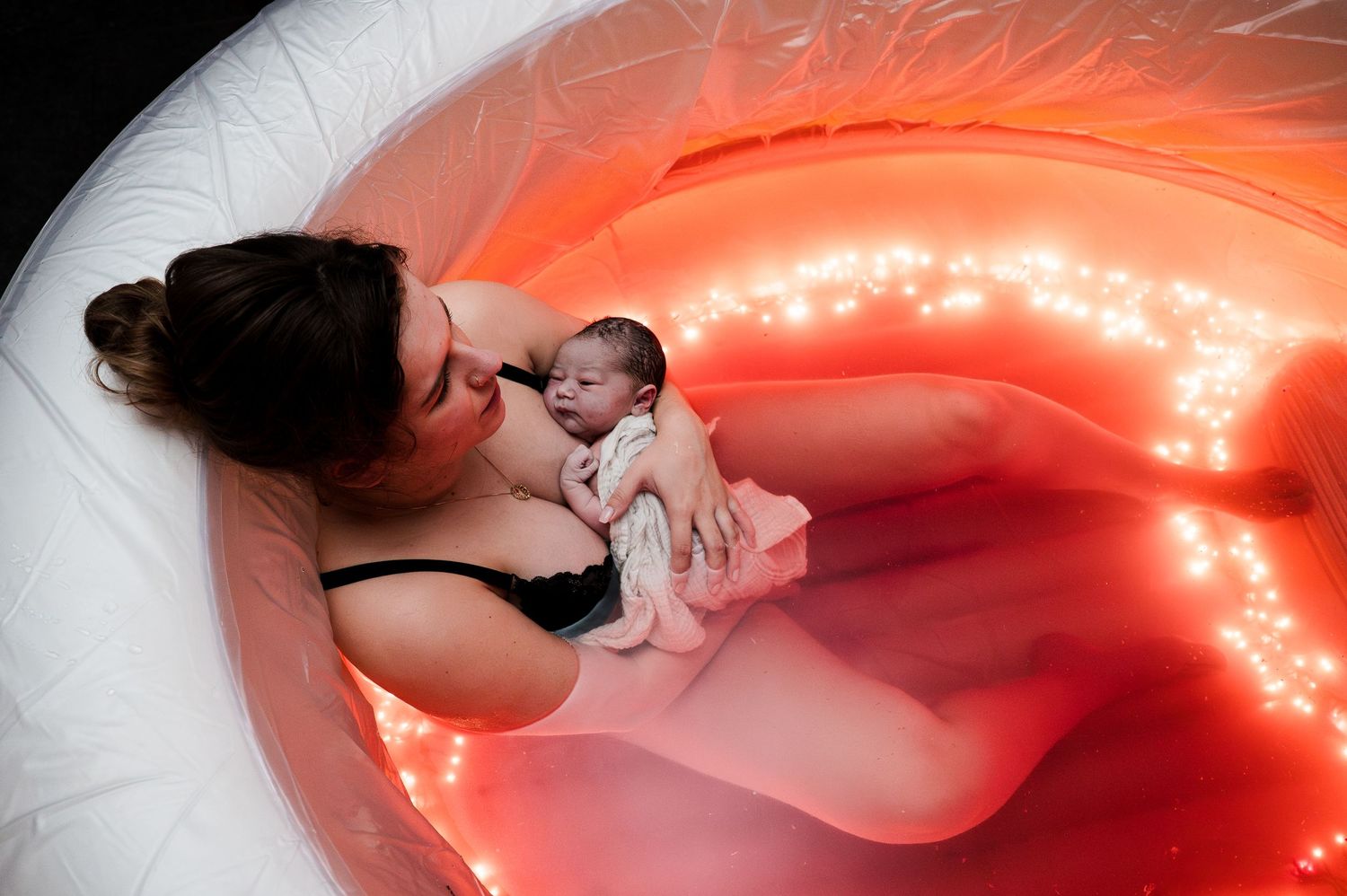 Mother holds newborn baby in glowing red water birth pool, sharing a tender moment after delivery.